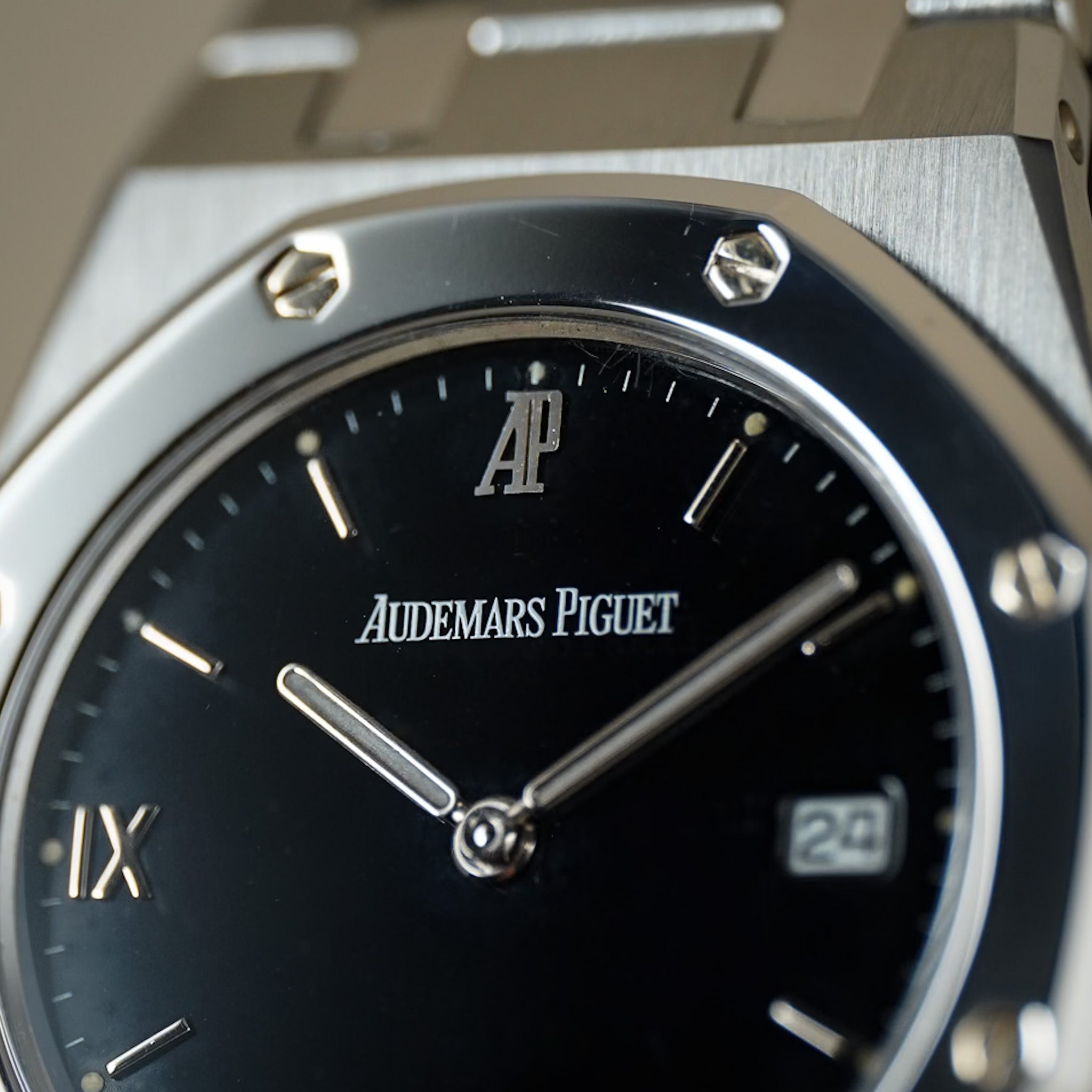 Close-up of an Audemars Piguet wristwatch with a black dial, silver-toned metal case, and visible AP logo at the 12 o'clock position.