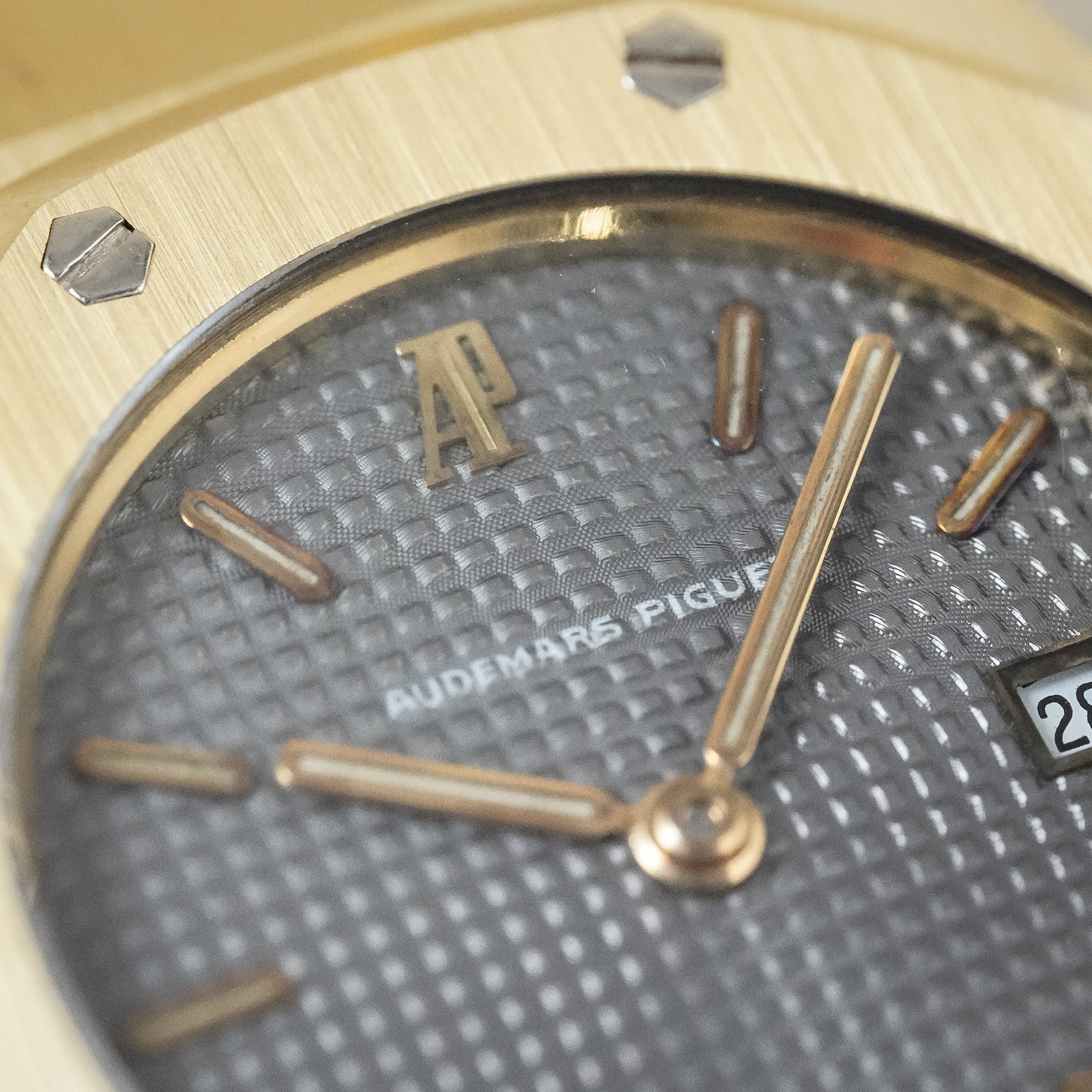 Close-up of an Audemars Piguet watch dial and bezel, showing a textured gray dial with gold-tone hands, indices, and the Audemars Piguet logo, set against a brushed gold-toned case.