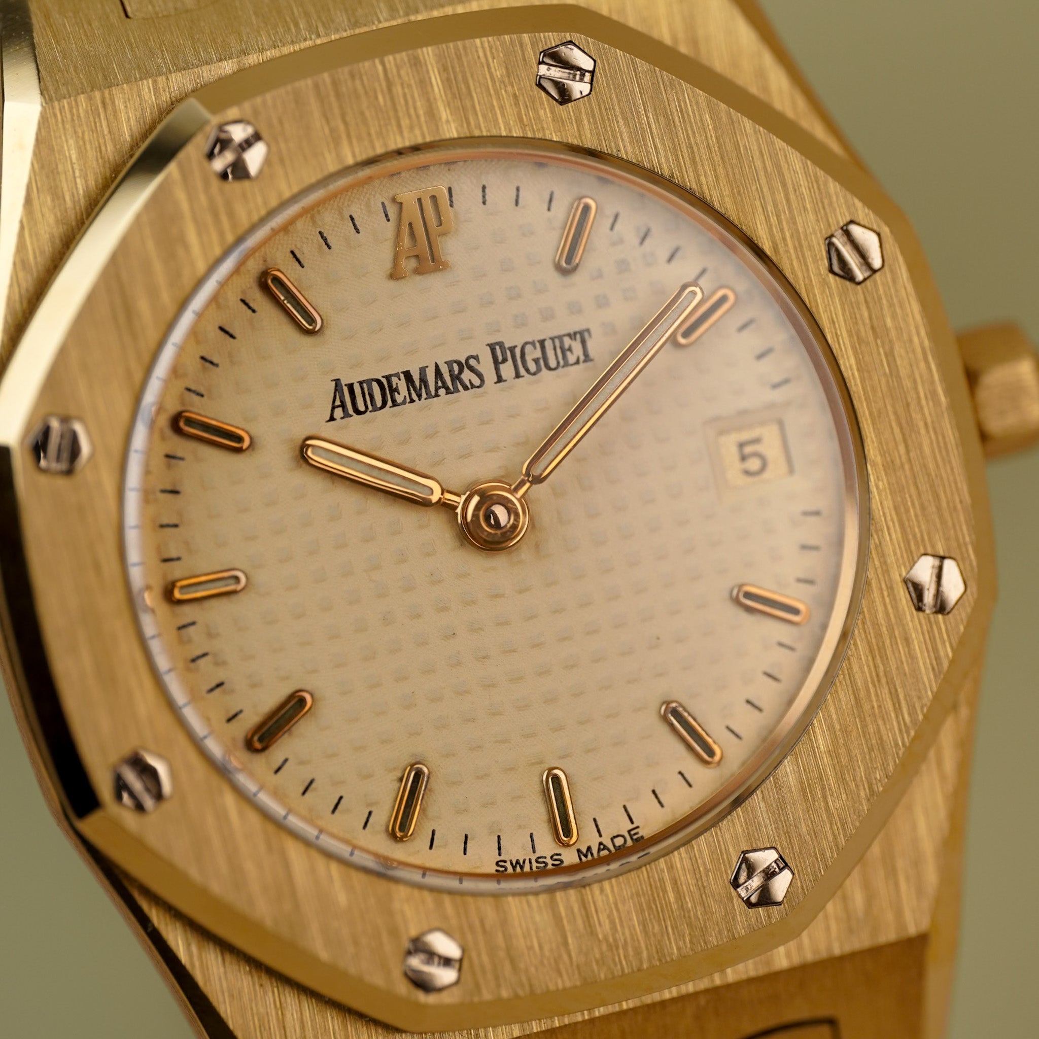 Closeup of a yellow gold Audemars Piguet Royal Oak ladies watch with a yellow gold octagonal bezel, yellow gold bracelet, a cream colored dial, and a date window at 3 o'clock, photographed on a light green background