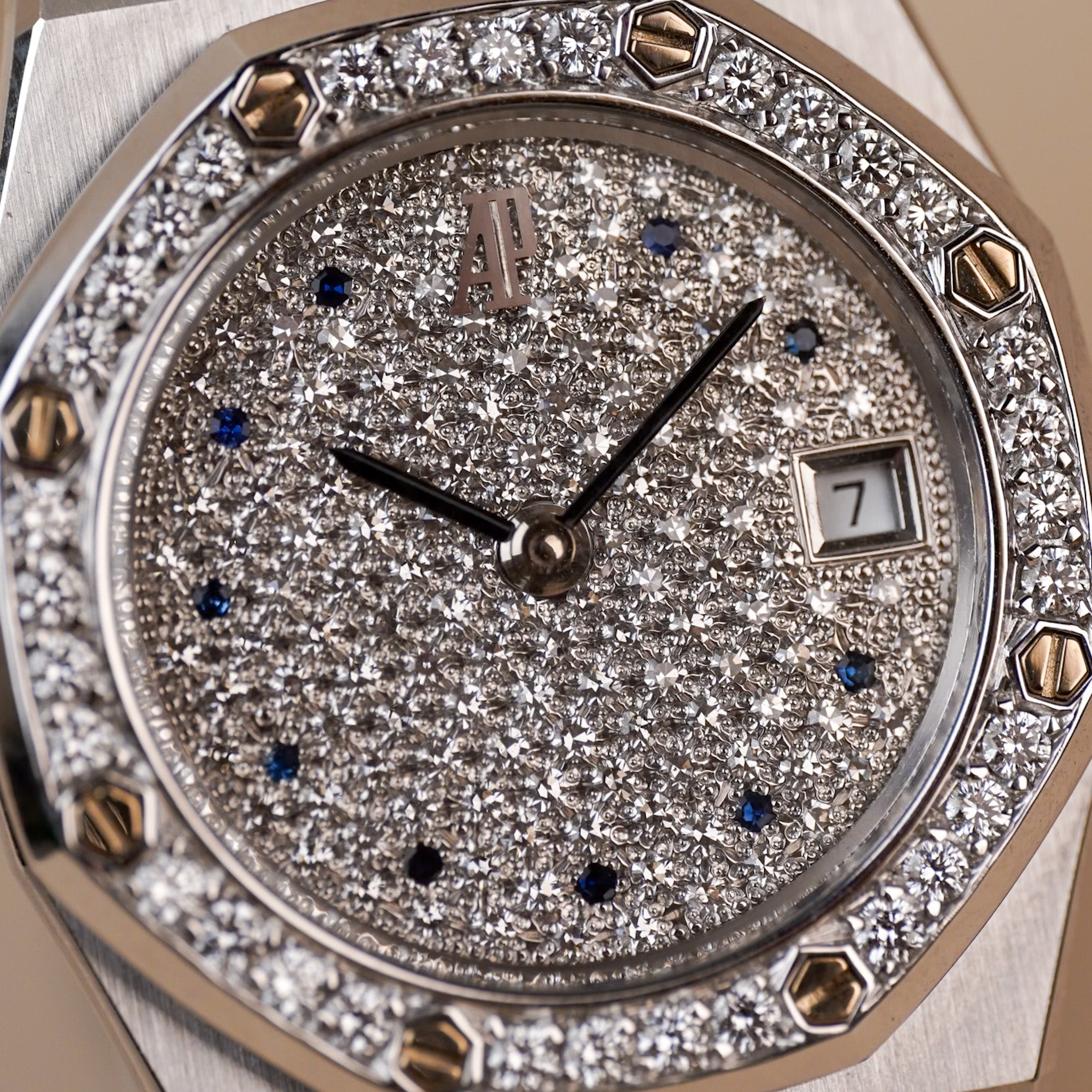 Close-up of a luxury watch dial and octagonal bezel, heavily set with sparkling pavé diamonds. The silver-toned case features gold-toned screw accents around the bezel, and the dial has black hands, blue sapphire markers, and a date window.