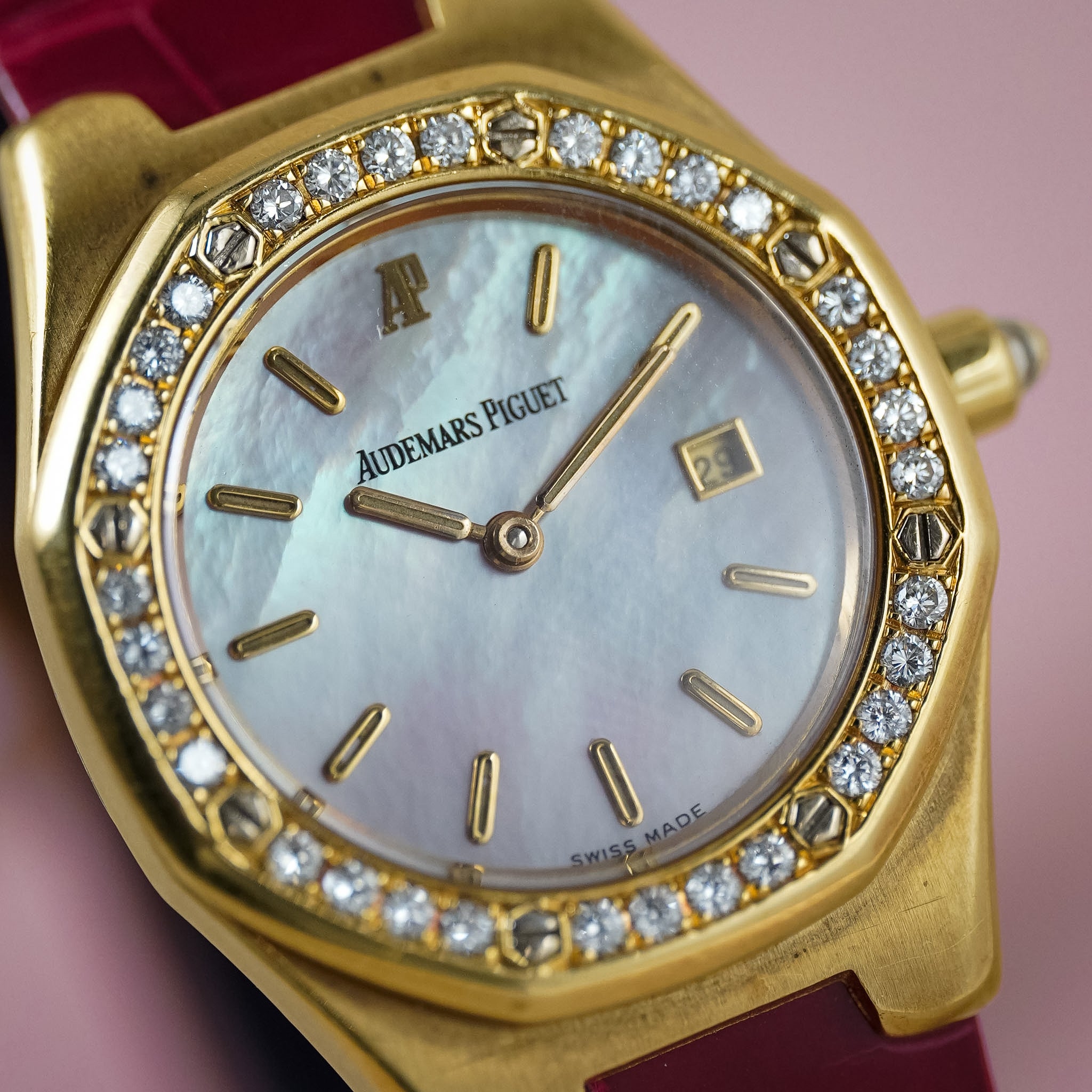 Audemars Piguet Royal Oak watch with a gold case, crystal-encrusted bezel, mother-of-pearl dial, and a deep red leather strap, shown in a close-up shot against a soft pink background.