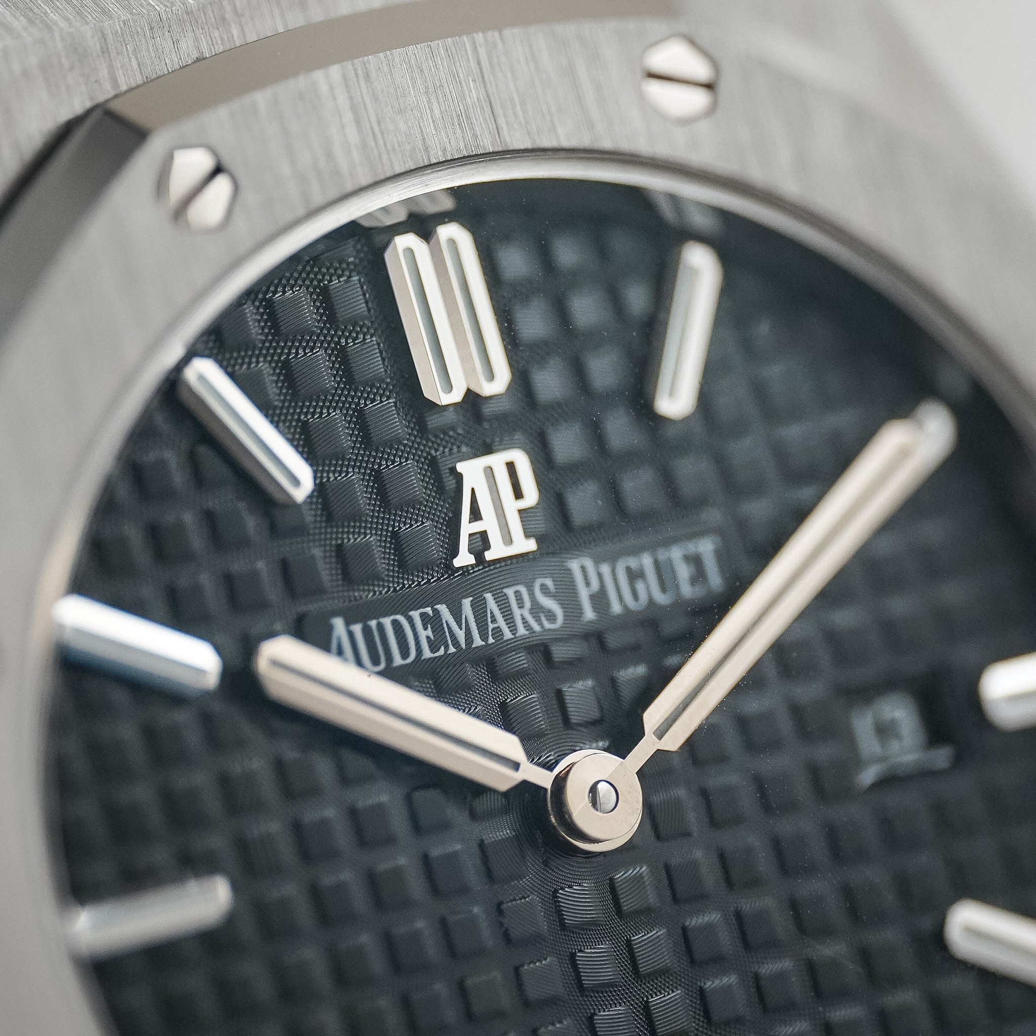 Close-up of an Audemars Piguet watch dial and bezel, showing a textured black dial with silver-tone hands, baton indices, and the AP logo.