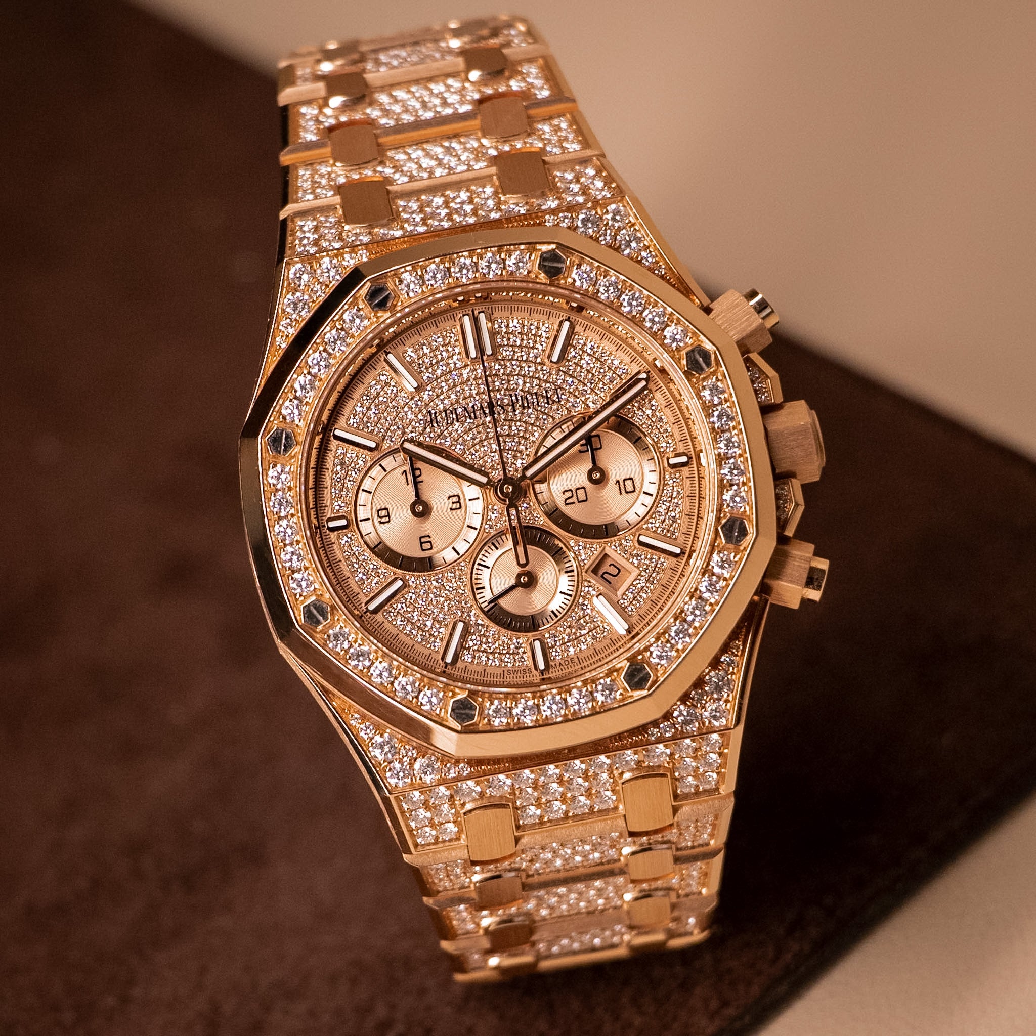 A luxury rose gold chronograph watch with a diamond-encrusted octagonal bezel and matching diamond-embellished bracelet, resting on a dark brown surface.