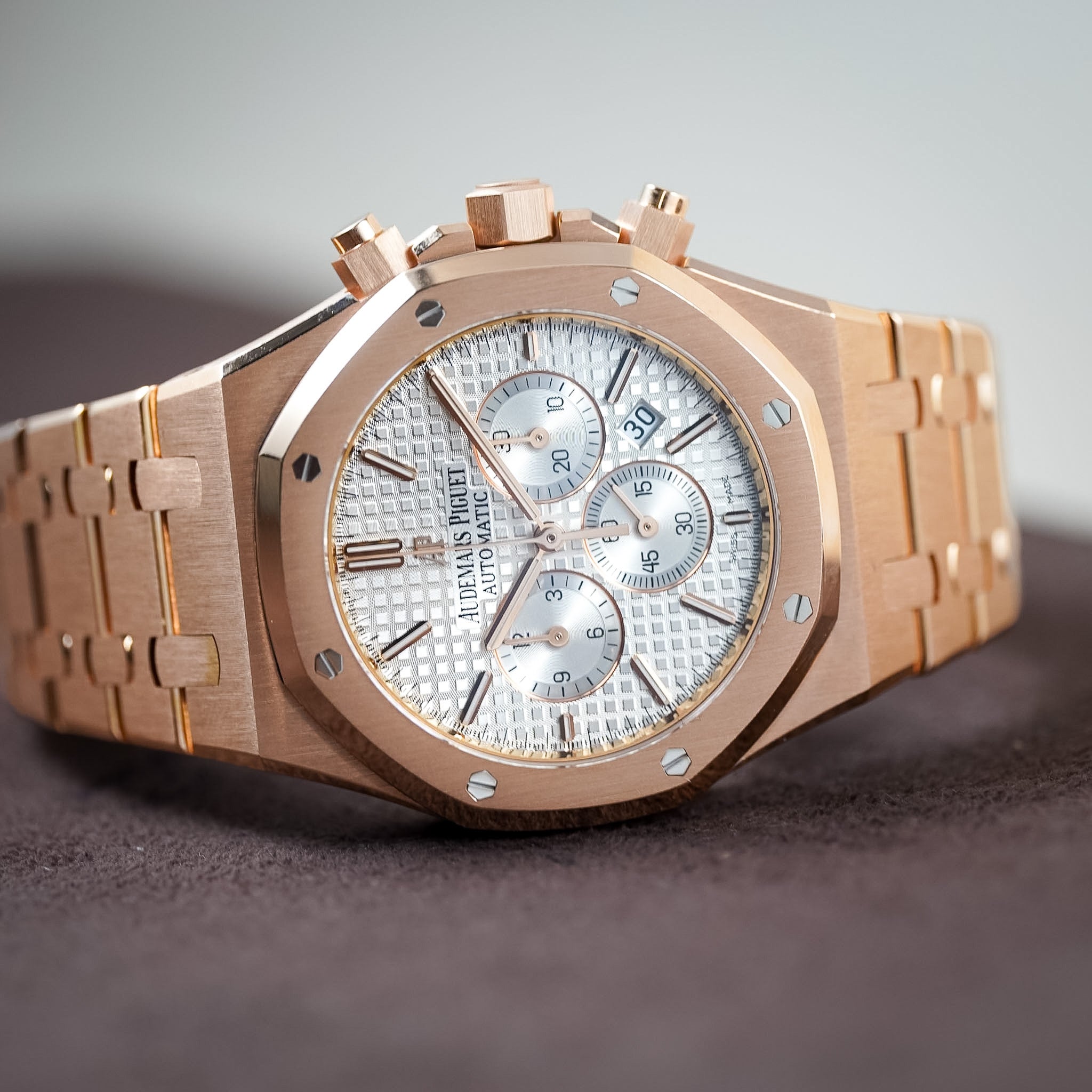 A close-up shot of a luxury watch with a rose gold case and bracelet, featuring a textured silver-white dial with three sub-dials. The brand name 'Audemars Piguet' is visible on the dial.