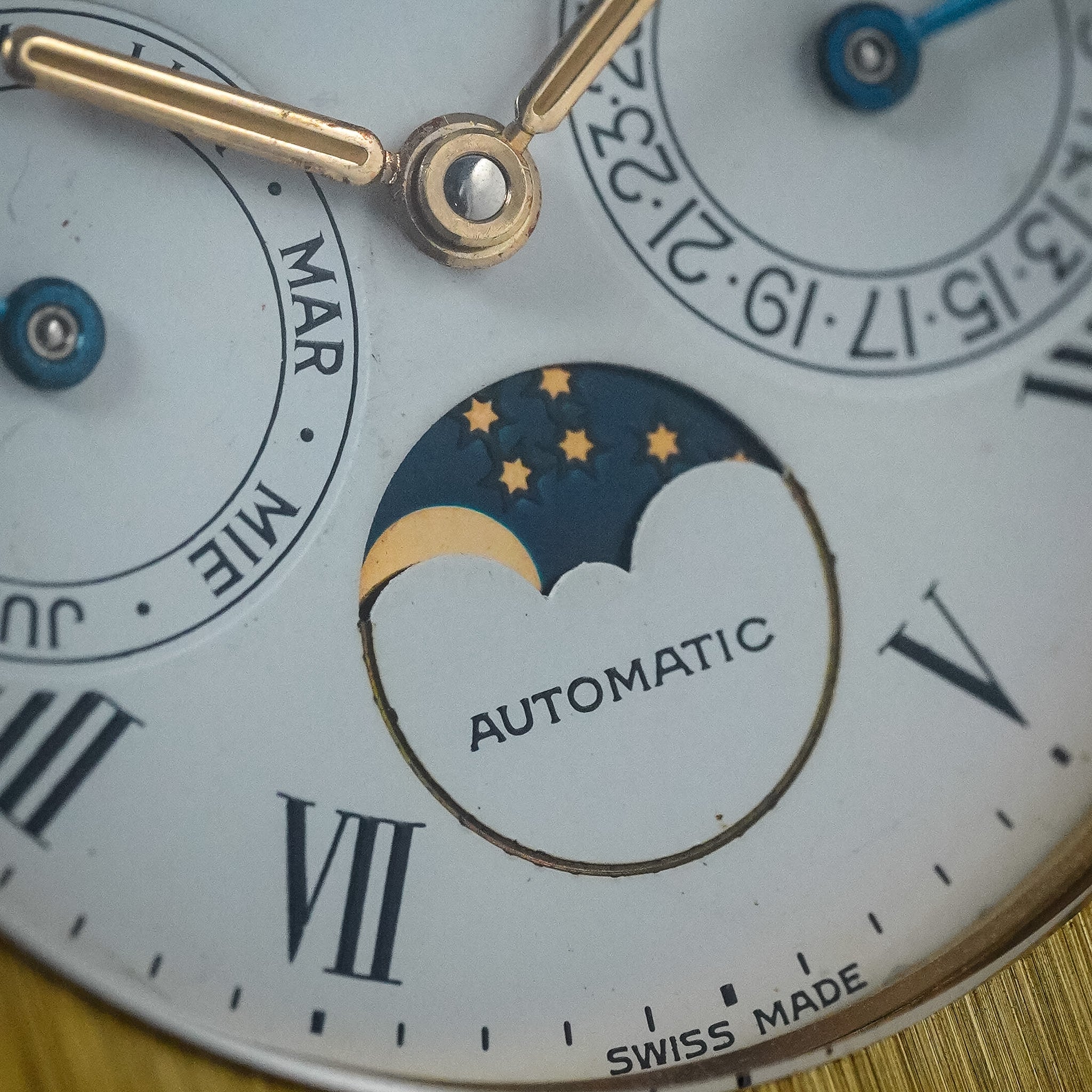 Close-up of a Swiss automatic watch dial with Roman numerals, a moonphase indicator with stars and crescent moon, gold-toned hands, and blue crown jewels on a pale blue face.