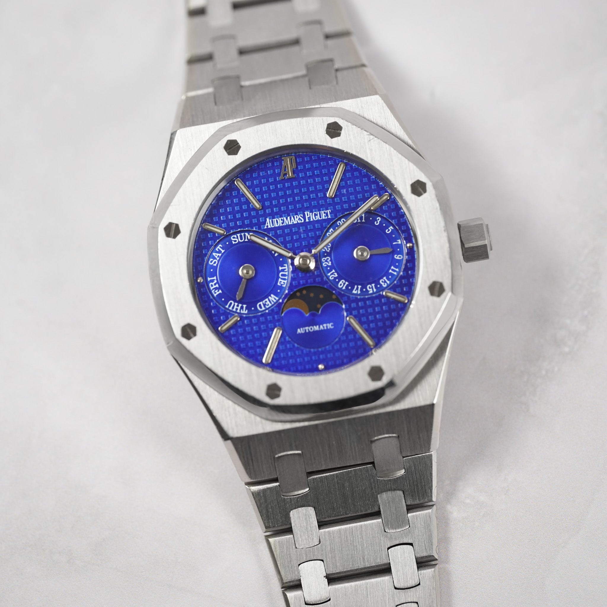 A silver-toned stainless steel wristwatch with an octagonal bezel and a vibrant blue dial featuring multiple sub-dials and a moon phase indicator. The brand name 'Audemars Piguet' is clearly visible on the dial.
