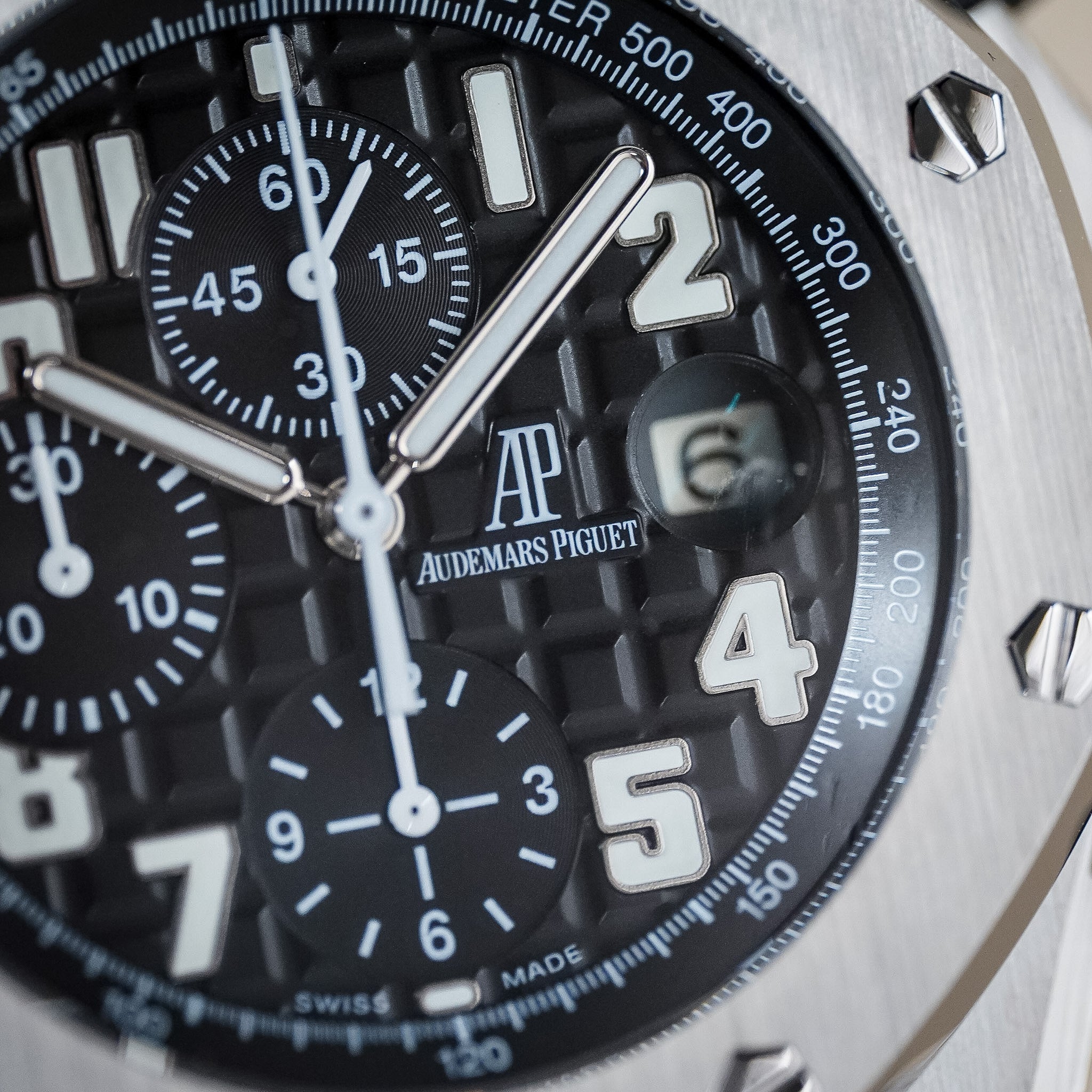 Closeup shot of a black Audemars Piguet wristwatch dial. It features three subdials, bold, white hour markers, and a date window. The watch has a steel bezel with exposed screws.