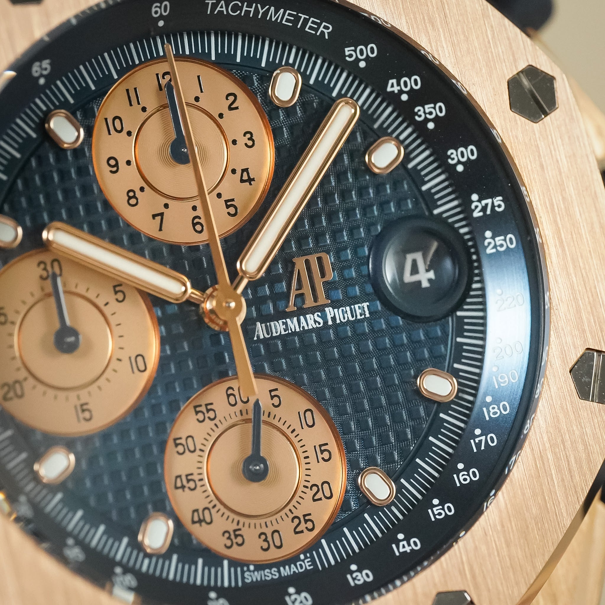 Close-up of an Audemars Piguet wristwatch dial and bezel, showing a textured dark blue chronograph face with rose gold subdials and hands, plus a black tachymeter scale.