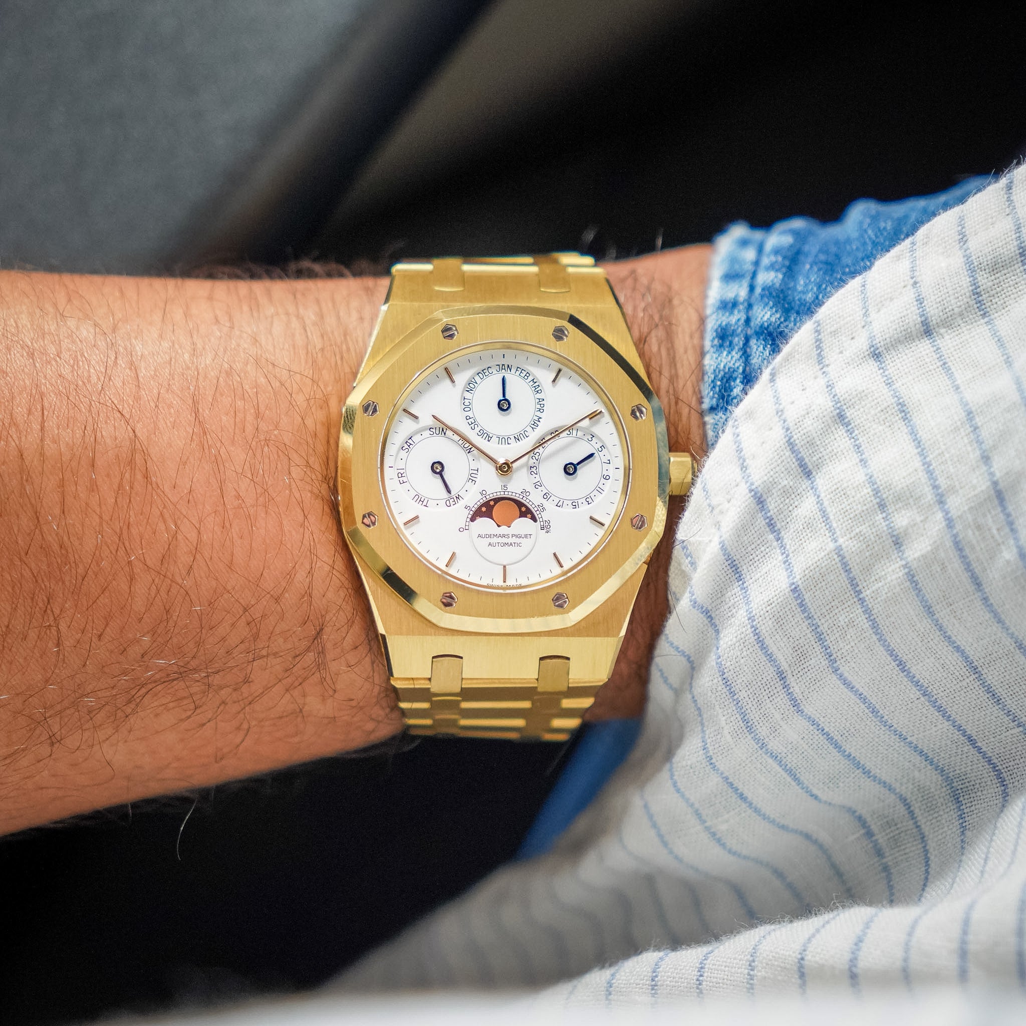 A gold-tone watch with a white dial and moonphase indicator, worn on a person's wrist next to a blue and white striped shirt.