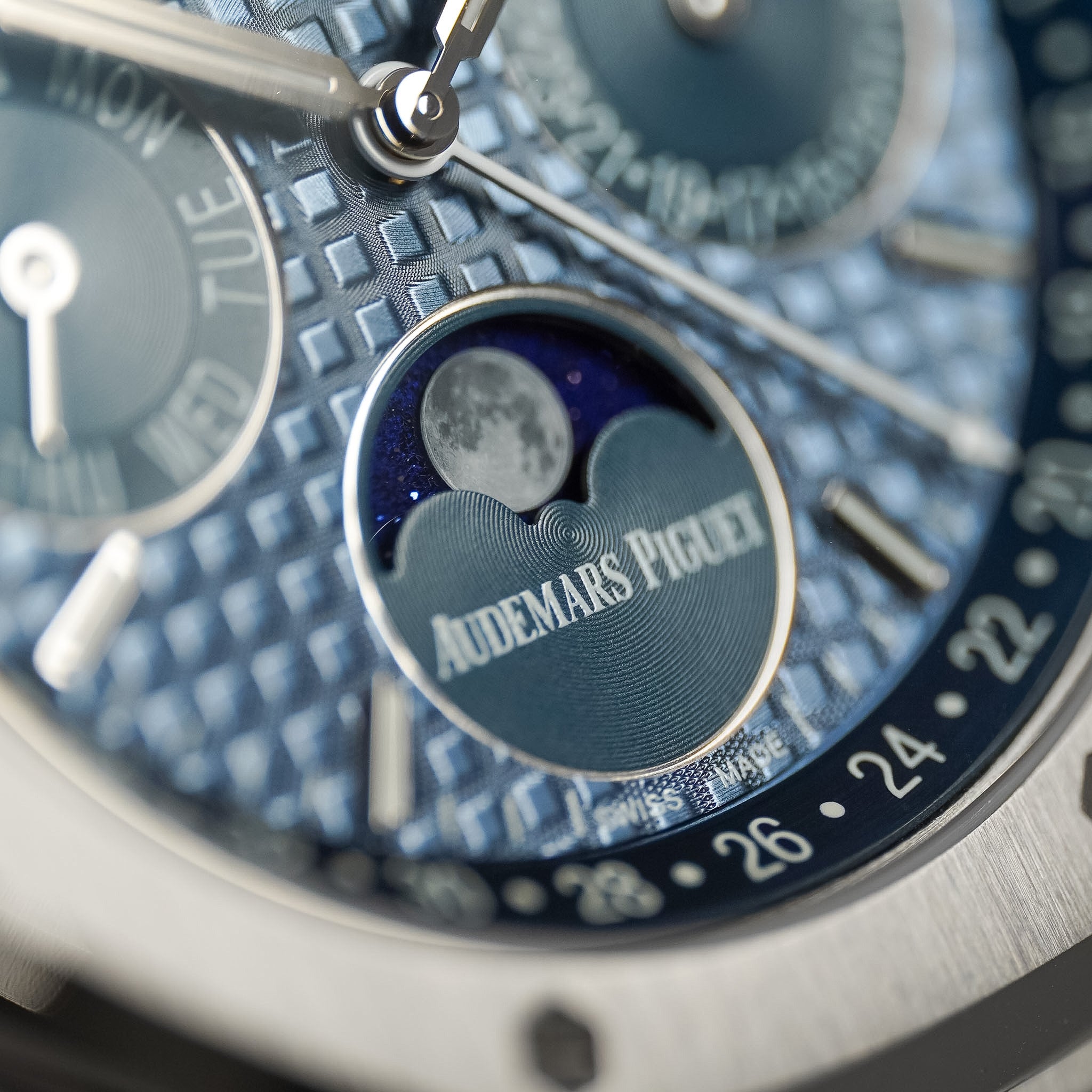 Close-up of an Audemars Piguet watch dial featuring a textured blue patterned case, silver hands and markers, and a moonphase subdial window with the brand name visible.