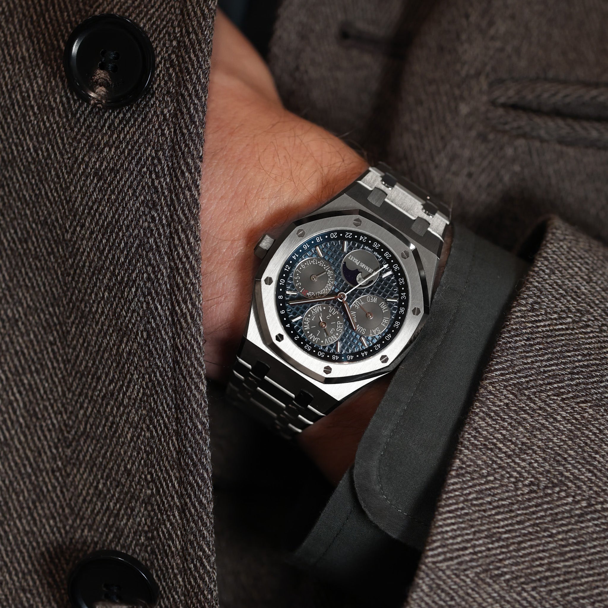 A man's wrist wearing a steel perpetual calendar wristwatch, reaching into his grey herringbone jacket.