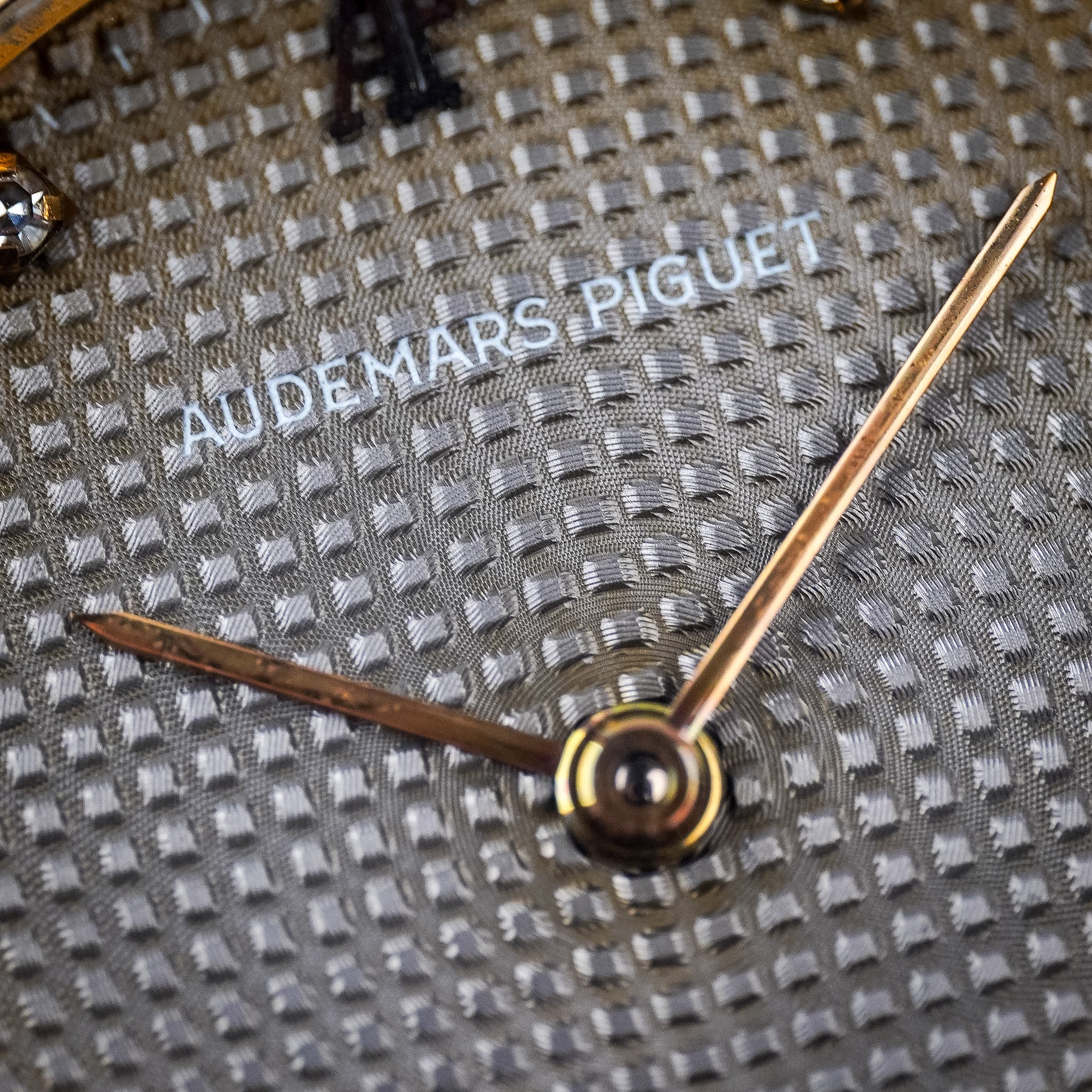 Closeup of an Audemars Piguet Royal Oak dial. It's grey in color with a geometric pattern imprinted into the dial. You also see gold hands and the Audemars Piguet logo.