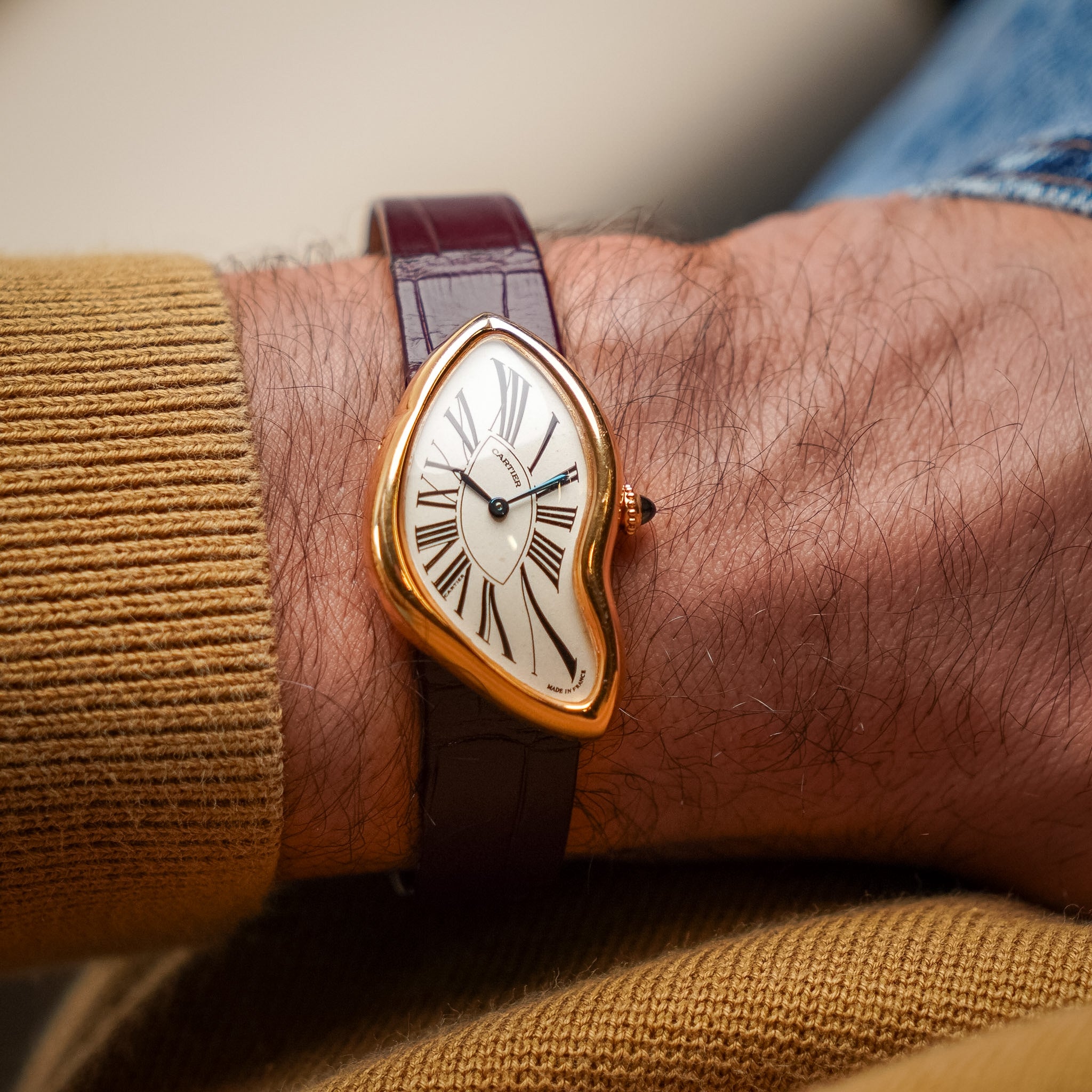 A close-up of a person's wrist wearing a unique asymmetrical gold-tone watch with a white dial, Roman numerals, and a dark brown leather strap.