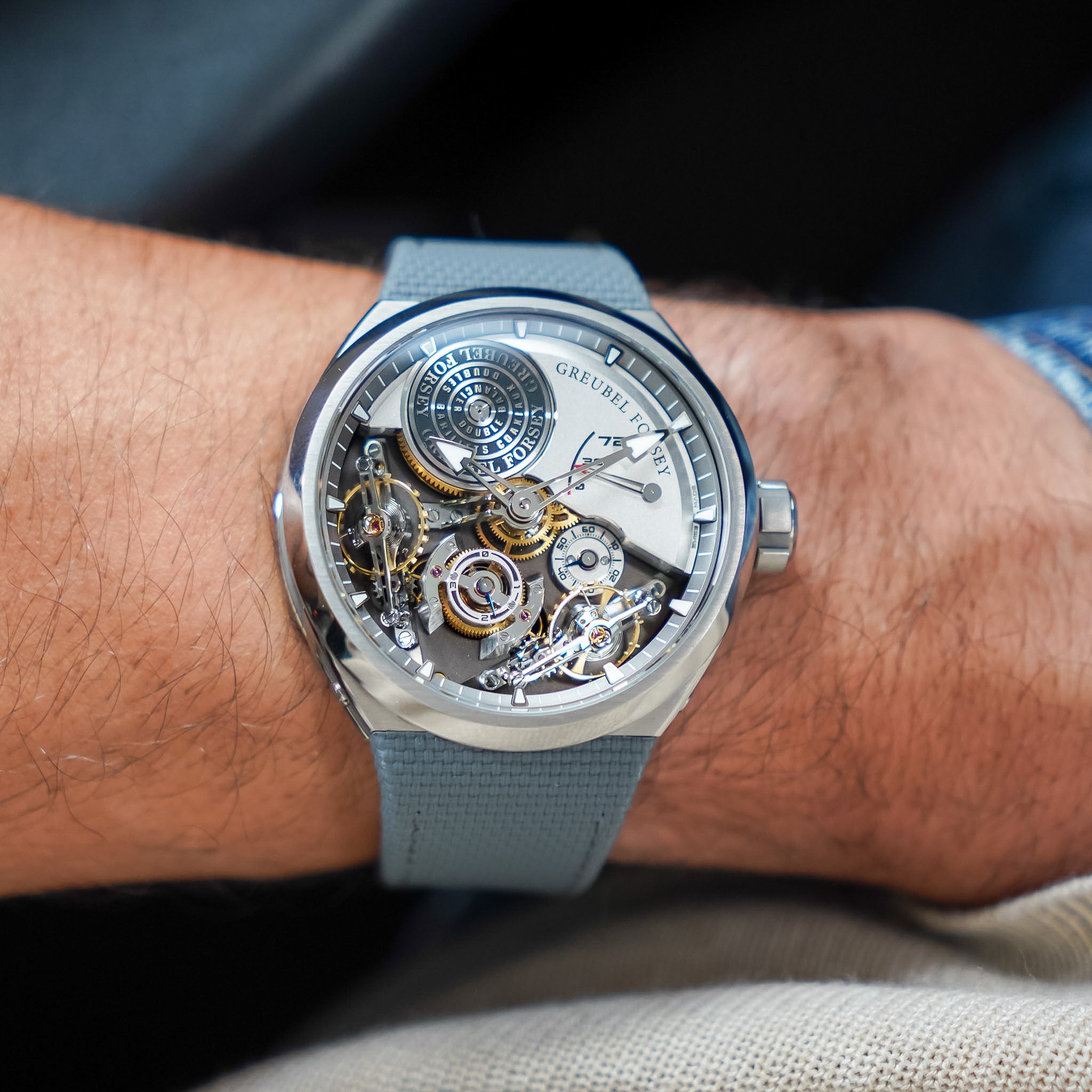 A close-up of a wrist wearing a skeleton watch with a gray textured strap, silver-toned case, and visible mechanical gears inside a clear dial.