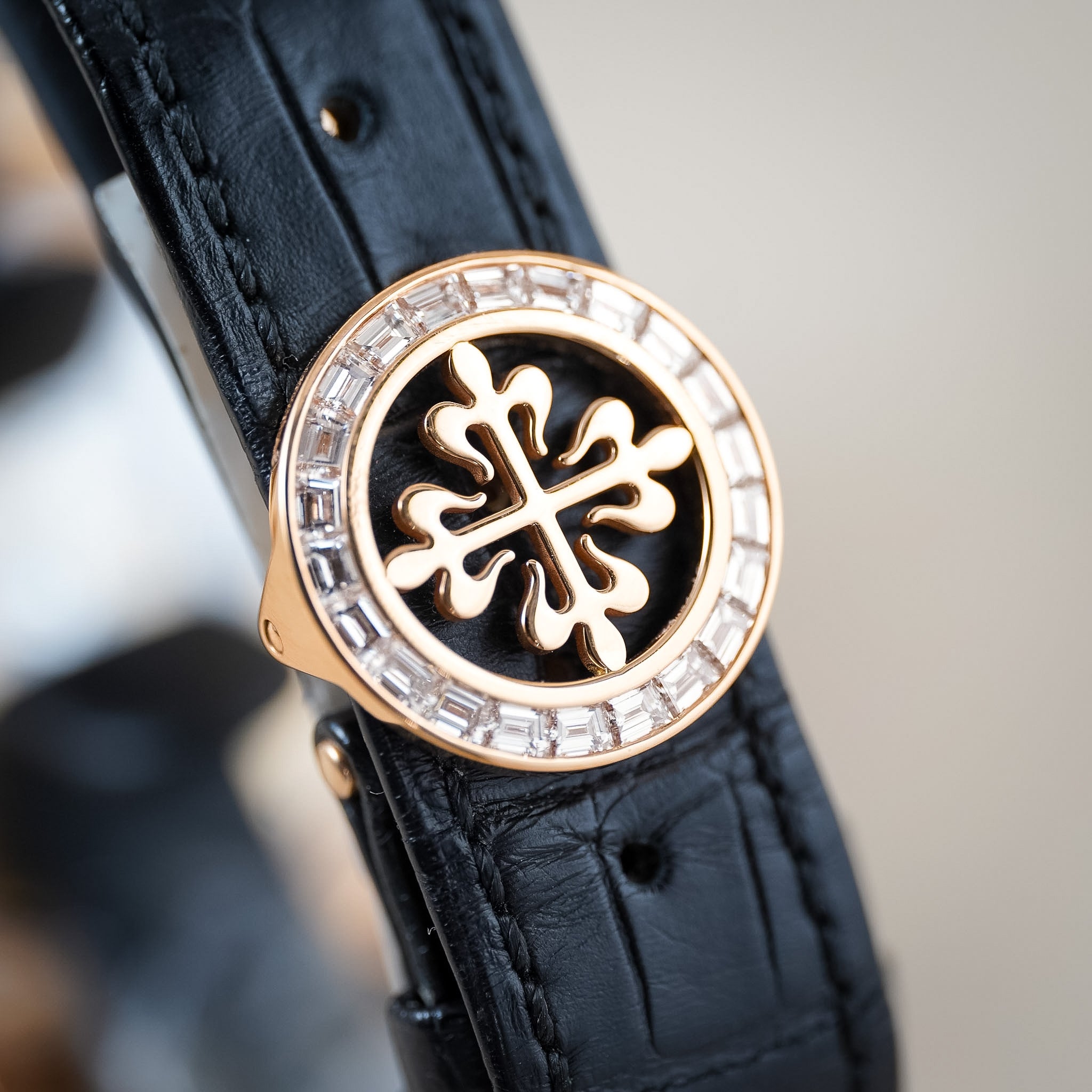 A close-up of a luxury watch featuring a black alligator leather strap and a round rose gold buckle adorned with clear crystals and a central black and rose gold emblem.