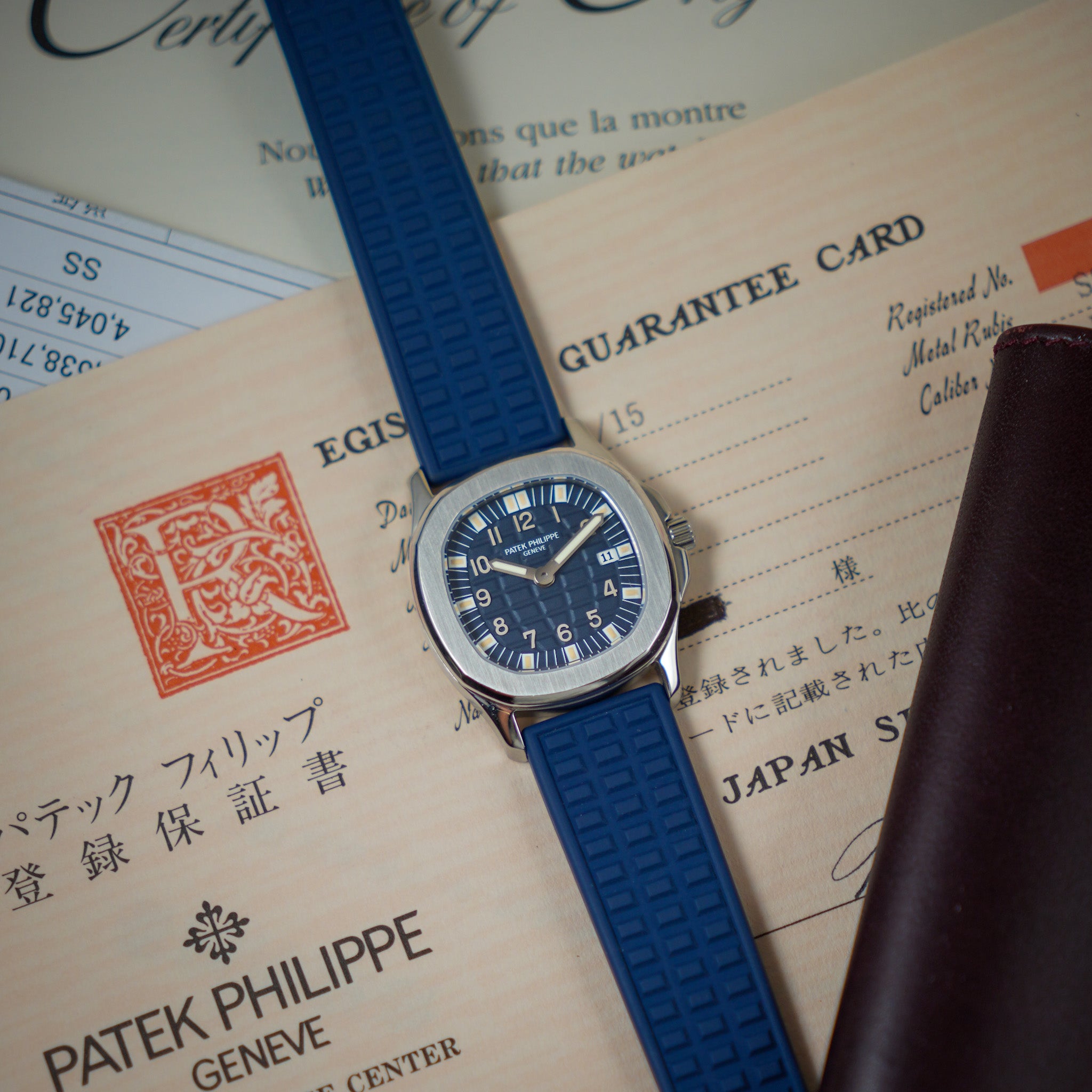 A Patek Philippe wristwatch with a blue textured rubber strap and a silver-toned stainless steel case, displayed diagonally on top of vintage Japanese guarantee documents next to a brown leather wallet.