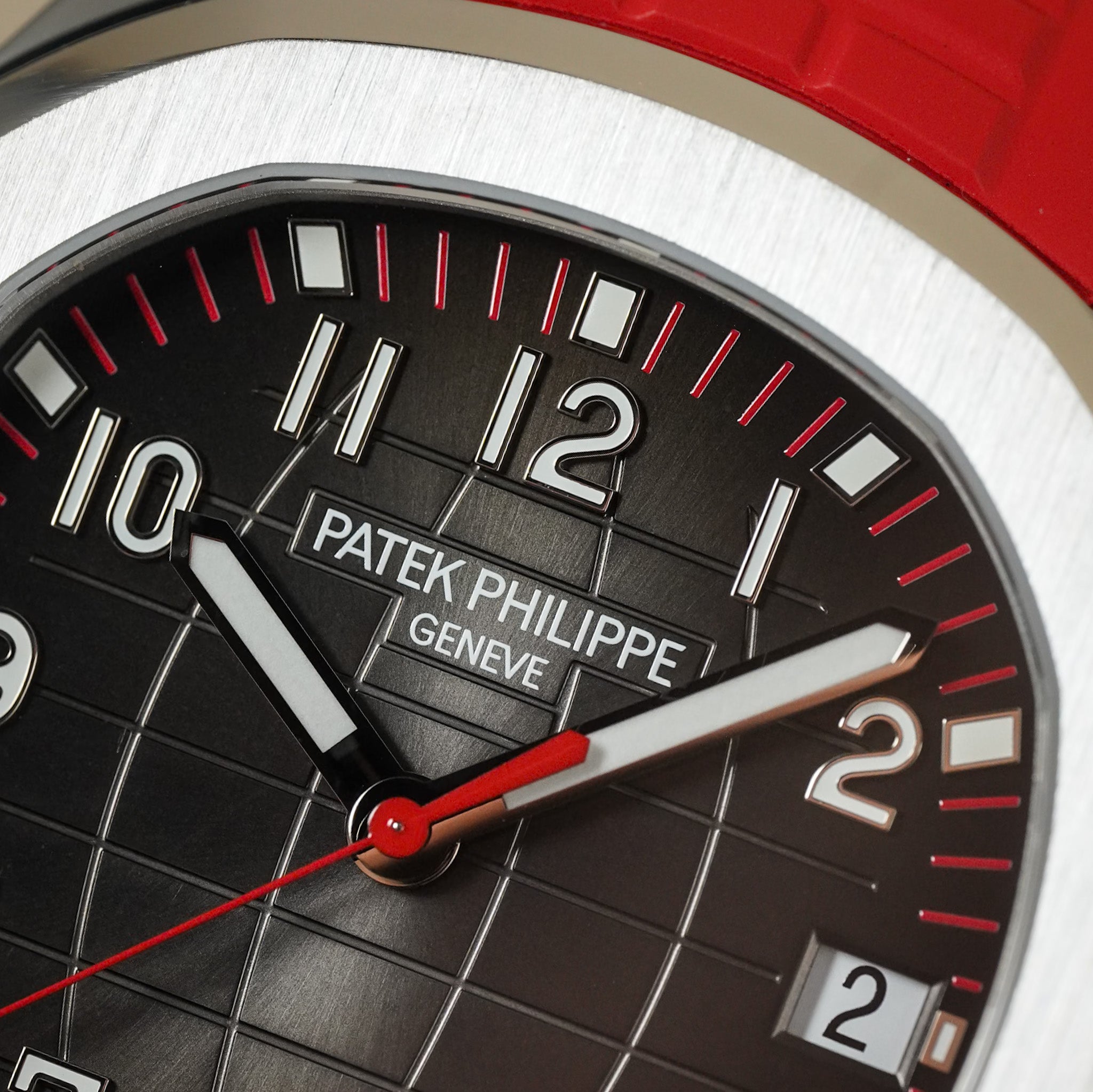 Close-up of a Patek Philippe wristwatch dial showing a black textured grid face, silver-tone numerals and hands, a red seconds hand, and part of a red strap against a brushed silver case.