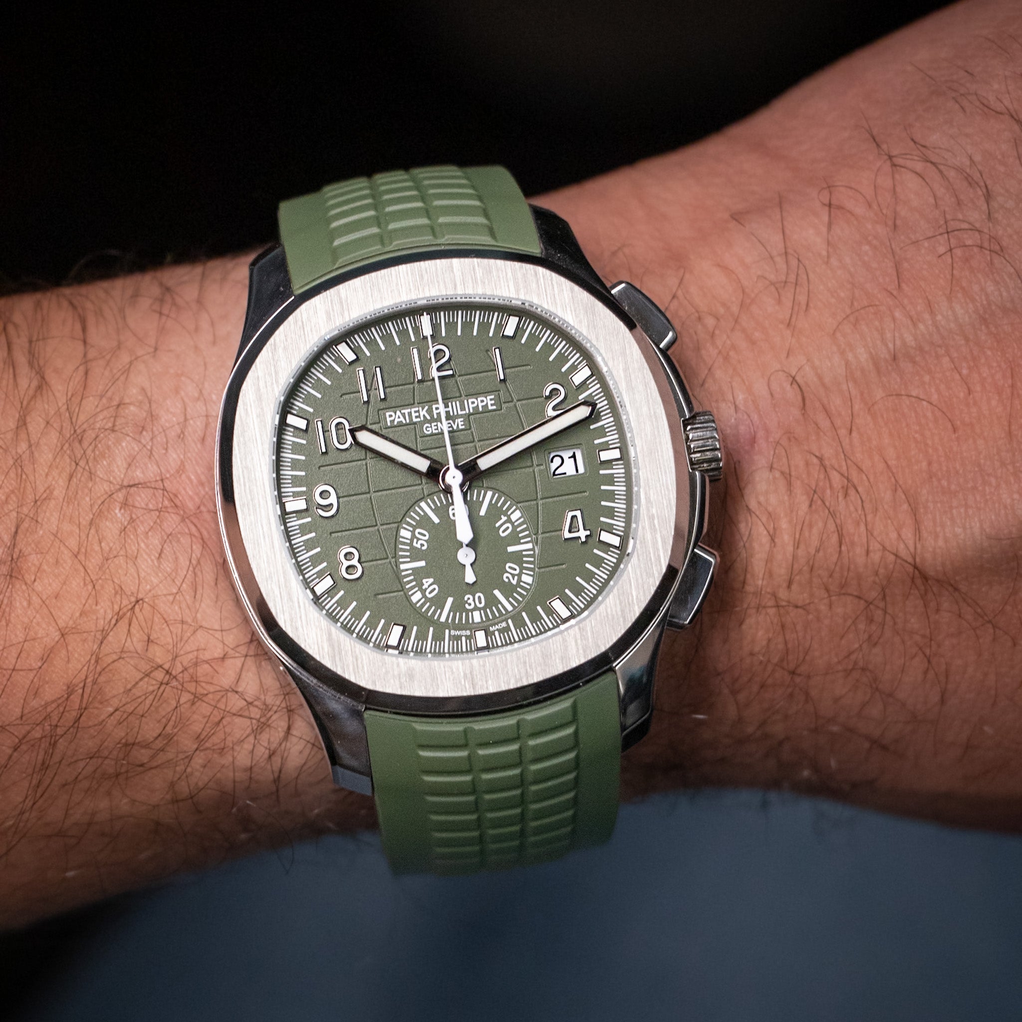 A wristwatch with an olive green rubber strap and a matching green dial, worn on a person's wrist. The watch has a silver-toned white gold case and a date window.