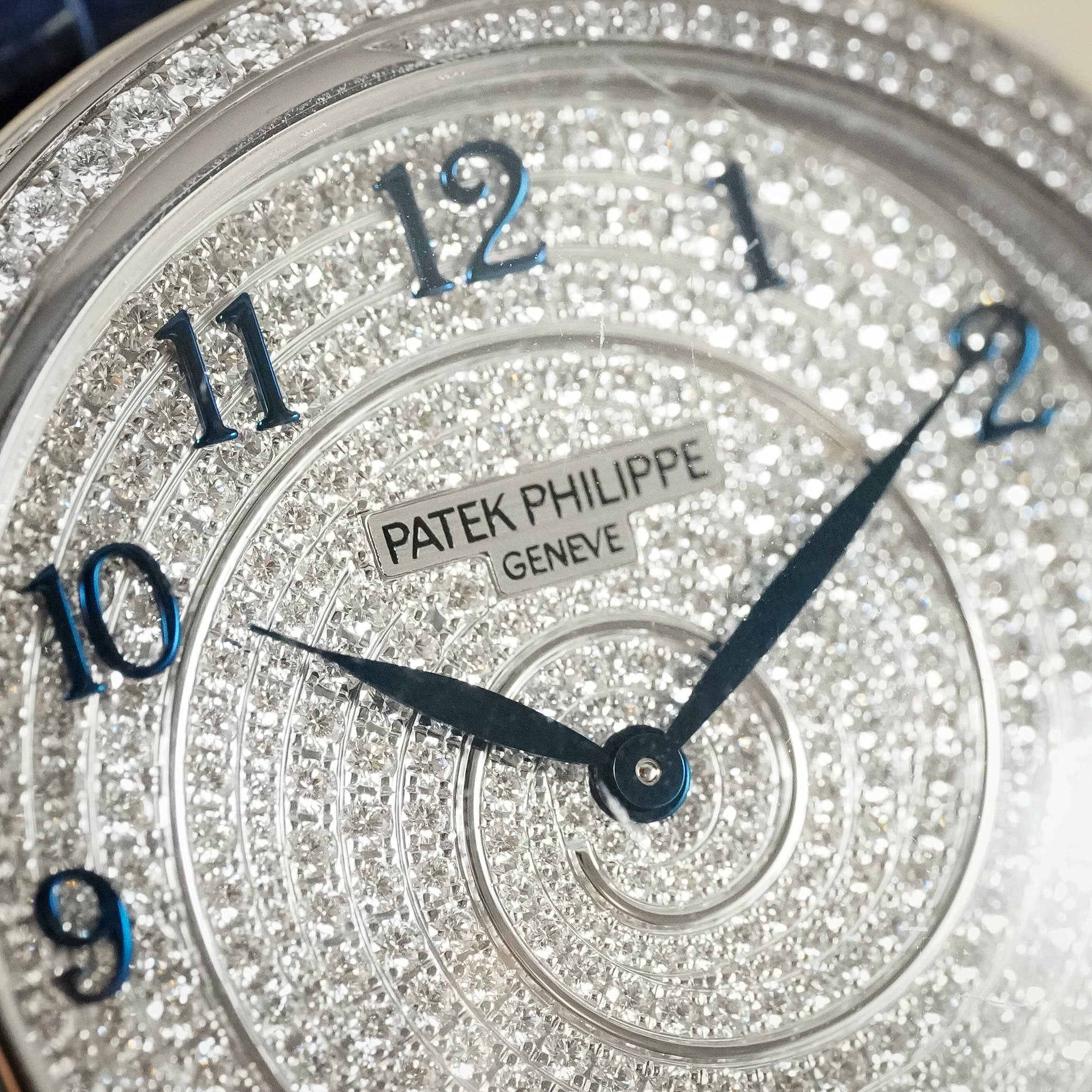 Close-up of a Patek Philippe watch dial covered in sparkling diamonds, with blue Arabic numerals and blue hands on a silver-toned case.