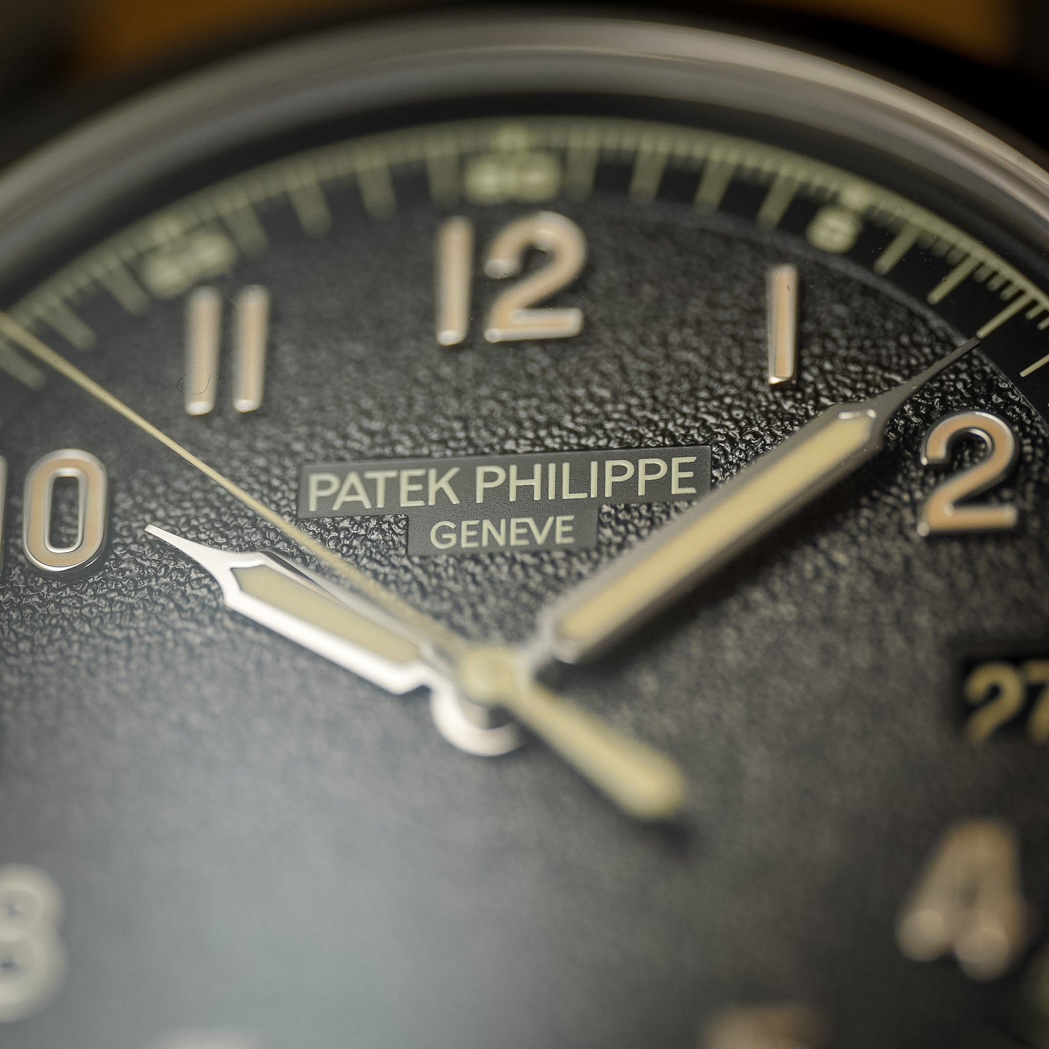 Close-up of a Patek Philippe watch dial with the logo in the top center. The dial is dark and textured with cream colored hands and hour markers.