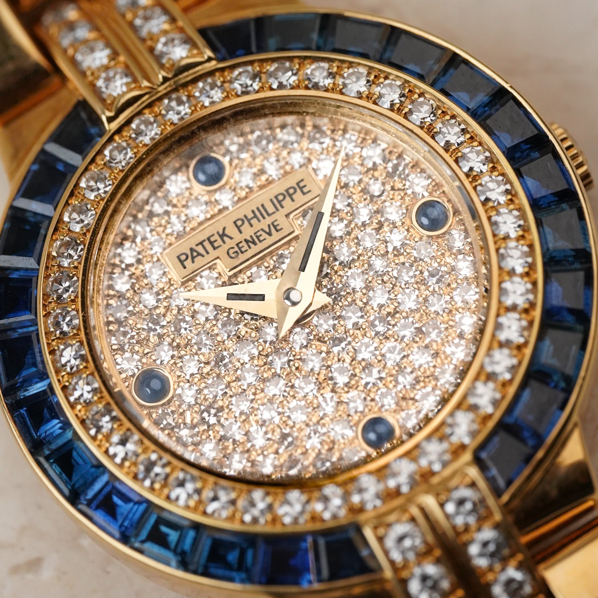 Close-up of a Patek Philippe Geneve watch with a gold-tone case, diamond-encrusted dial, and a blue bezel.