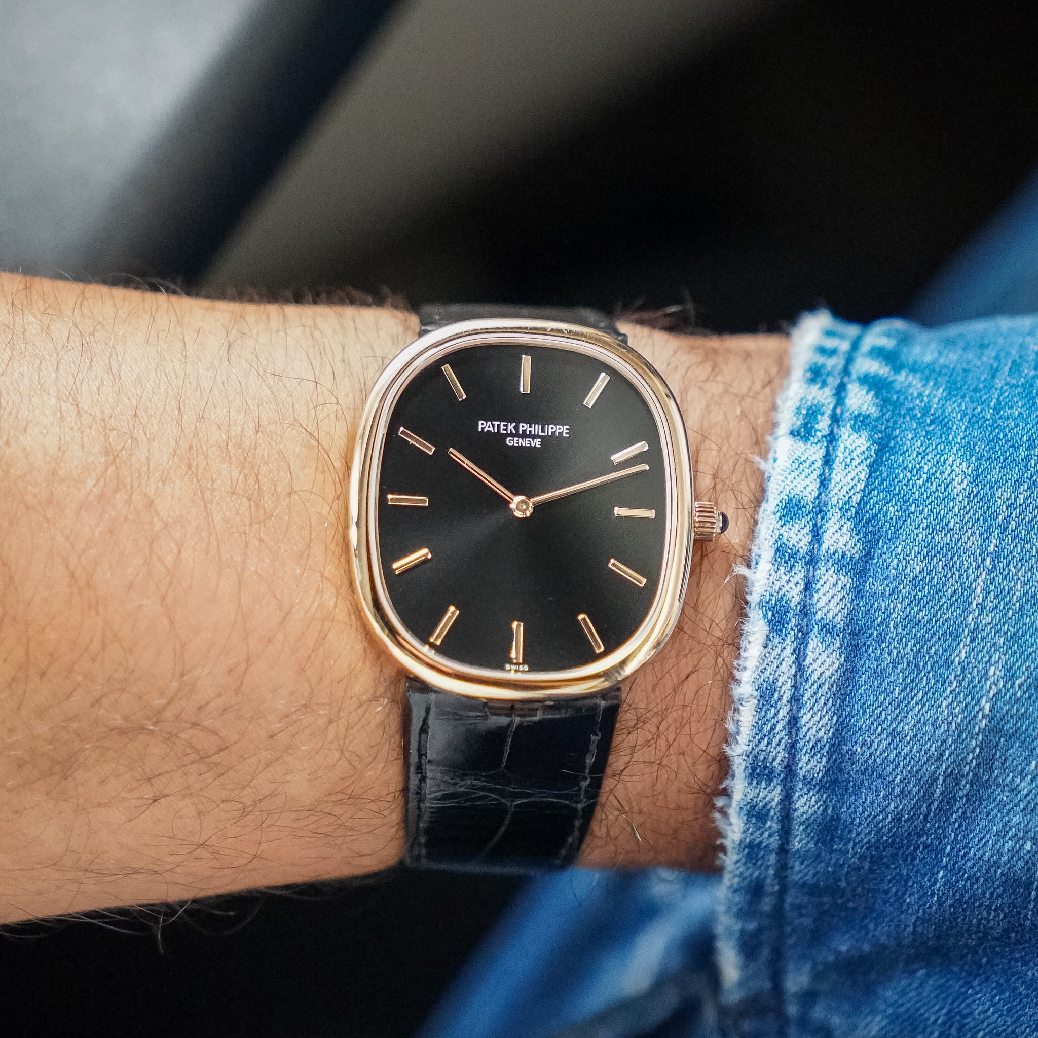 A close-up of a wrist wearing a Patek Philippe watch with a black dial, gold-tone case, and black leather strap, paired with blue denim clothing.