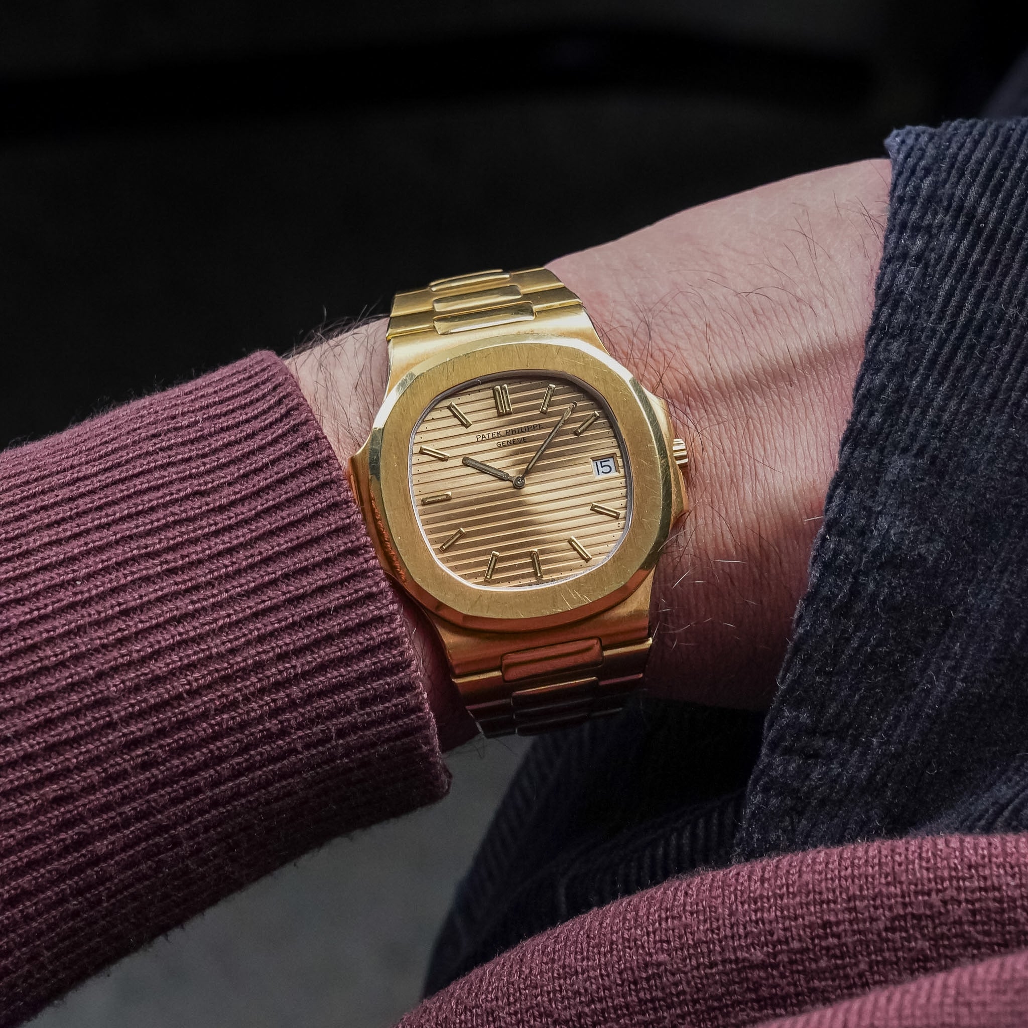 A yellow gold Patek Philippe Nautilus with a gold-tone dial, yellow gold case and bezel, gold hands and hour markers, and a date window at 3 o'clock. Shot on a man's wrist wearing a maroon sweater and black pants.