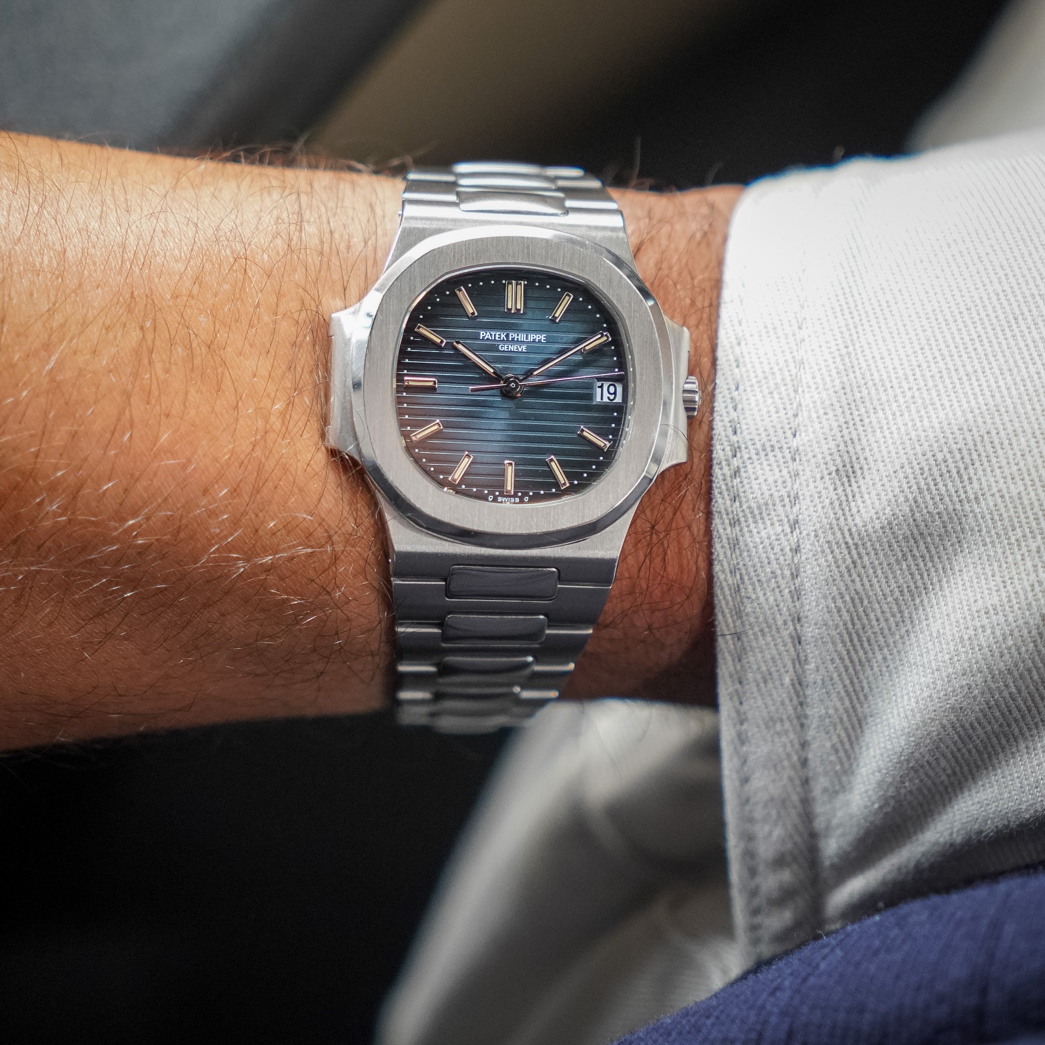 A stainless steel wristwatch with a blue sunburst dial and white gold indices, worn on a person's wrist with light gray pants.