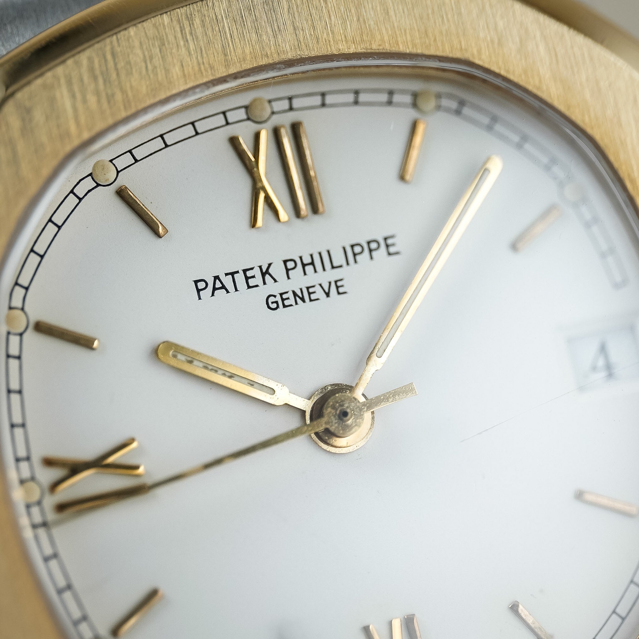 Close-up of a Patek Philippe Nautilus dial in white with a yellow gold bezel, hands, and hour markers. The watch features a date window at 3 o'clock.