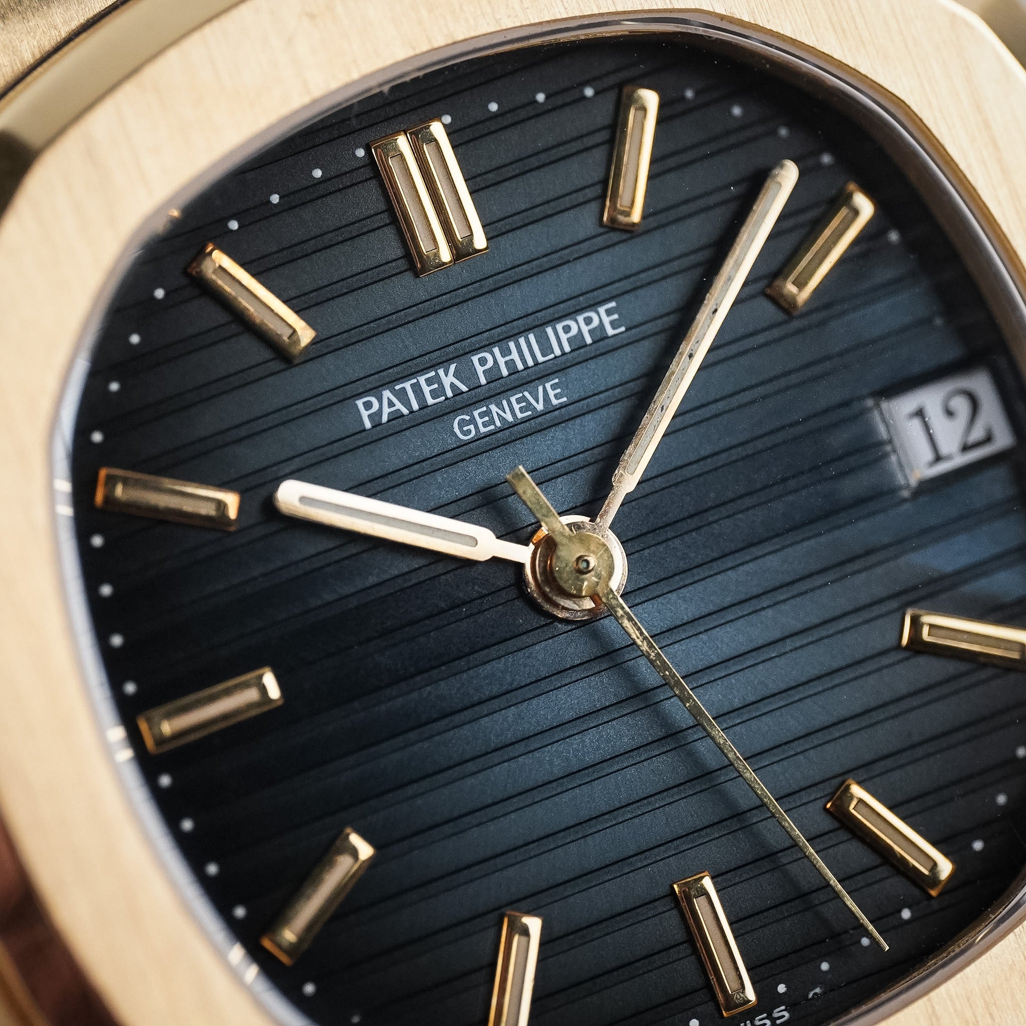 A close-up shot of a yellow gold Patek Philippe Nautilus with a ribbed blue dial, yellow gold hands and hour markers, and date window at 3 o'clock showing "12"