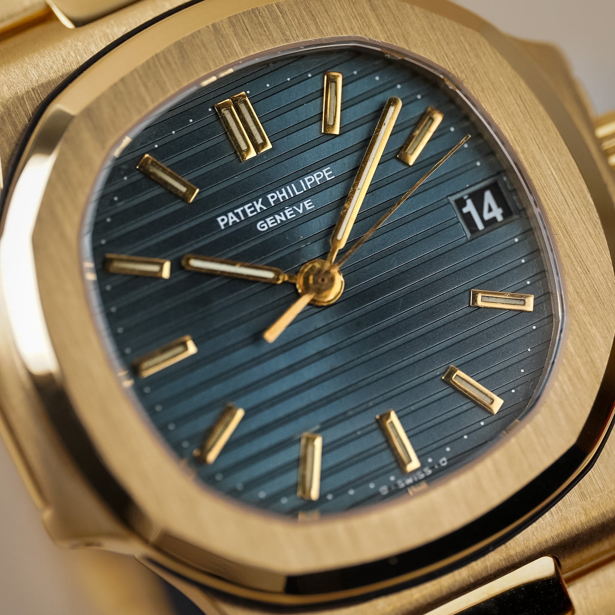 Closeup of a yellow gold Patek Philippe Nautilus with a textured blue dial, gold hands and hour markers, a gold bracelet, and a date window showing "14"