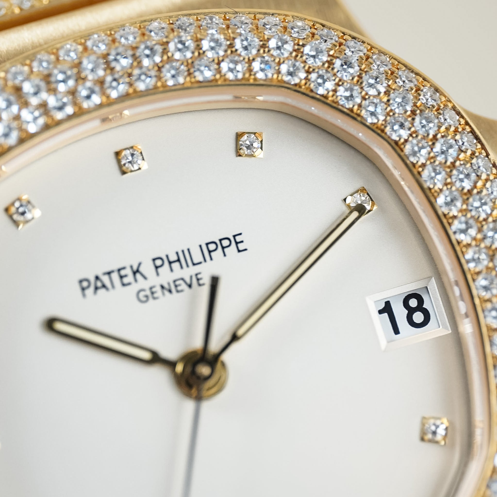 Close-up of a Patek Philippe watch face with a gold bezel set with diamonds, white dial, gold hands, and a date window showing 18.