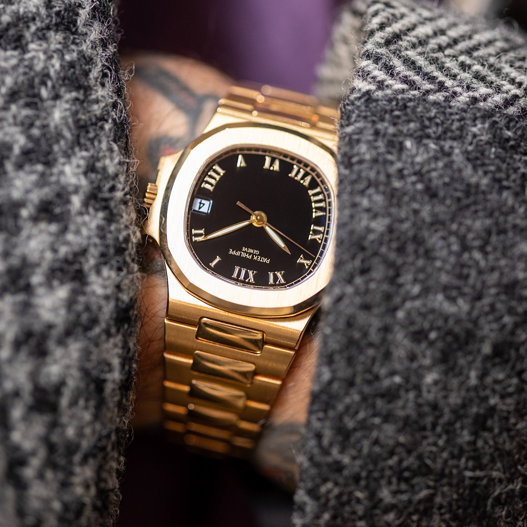 A close-up shot of a gold-tone wristwatch with a black dial and Roman numeral markers, worn on a person's wrist between gray textured knit sleeves.