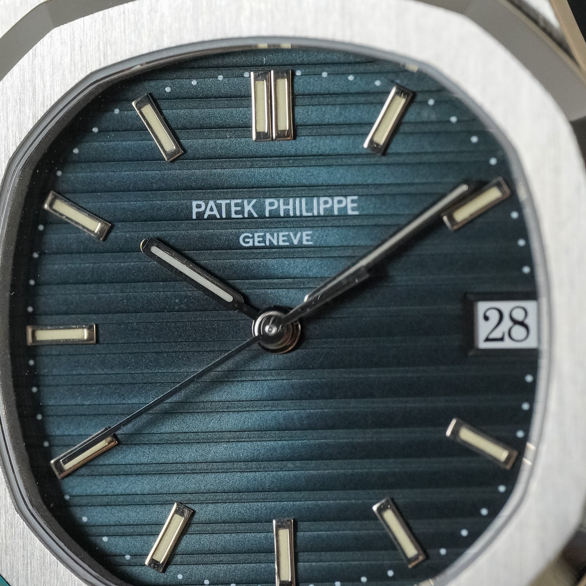 Close-up of a Patek Philippe wristwatch dial and bezel, showing a blue ribbed textured face with silver-tone baton markers and a date window displaying 28.