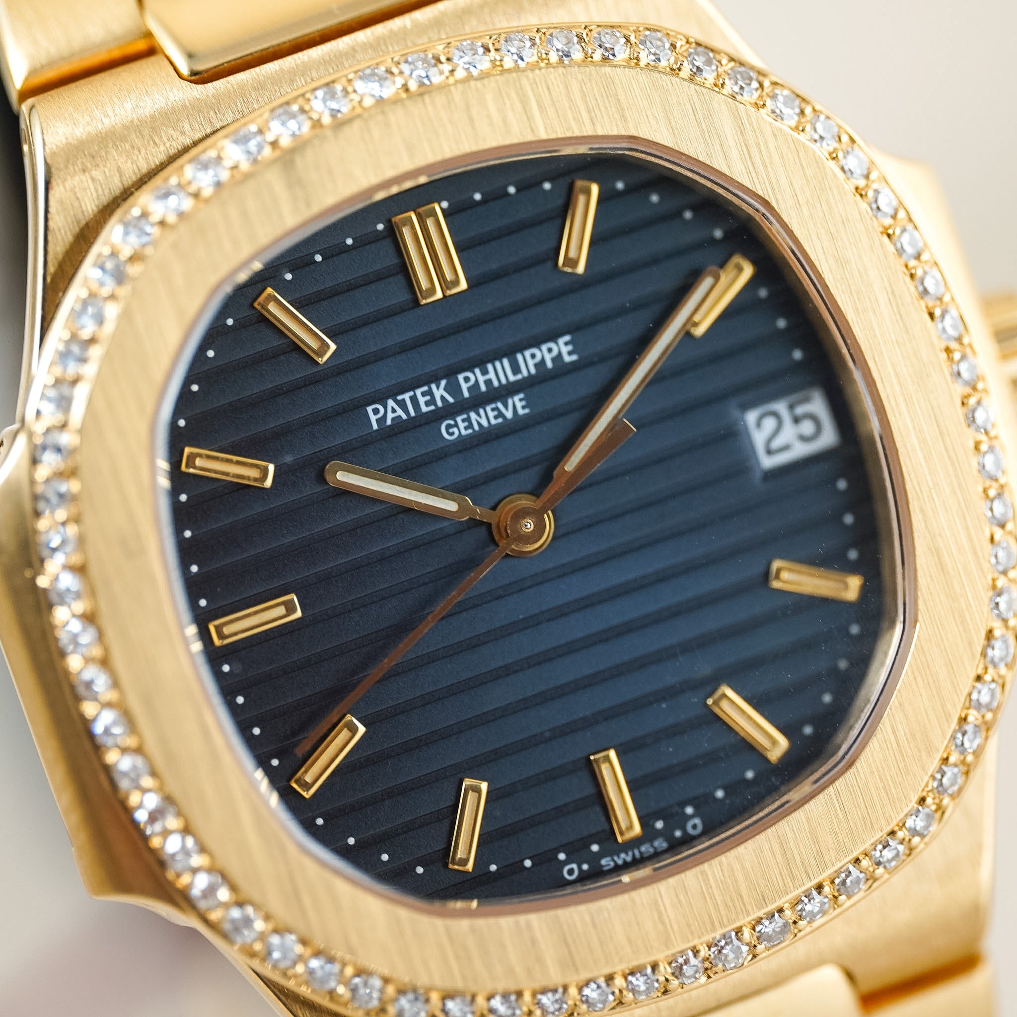 Close-up of a gold-tone Patek Philippe wristwatch with a dark blue striped dial, gold hour markers and hands, and a bezel set with small clear stones.