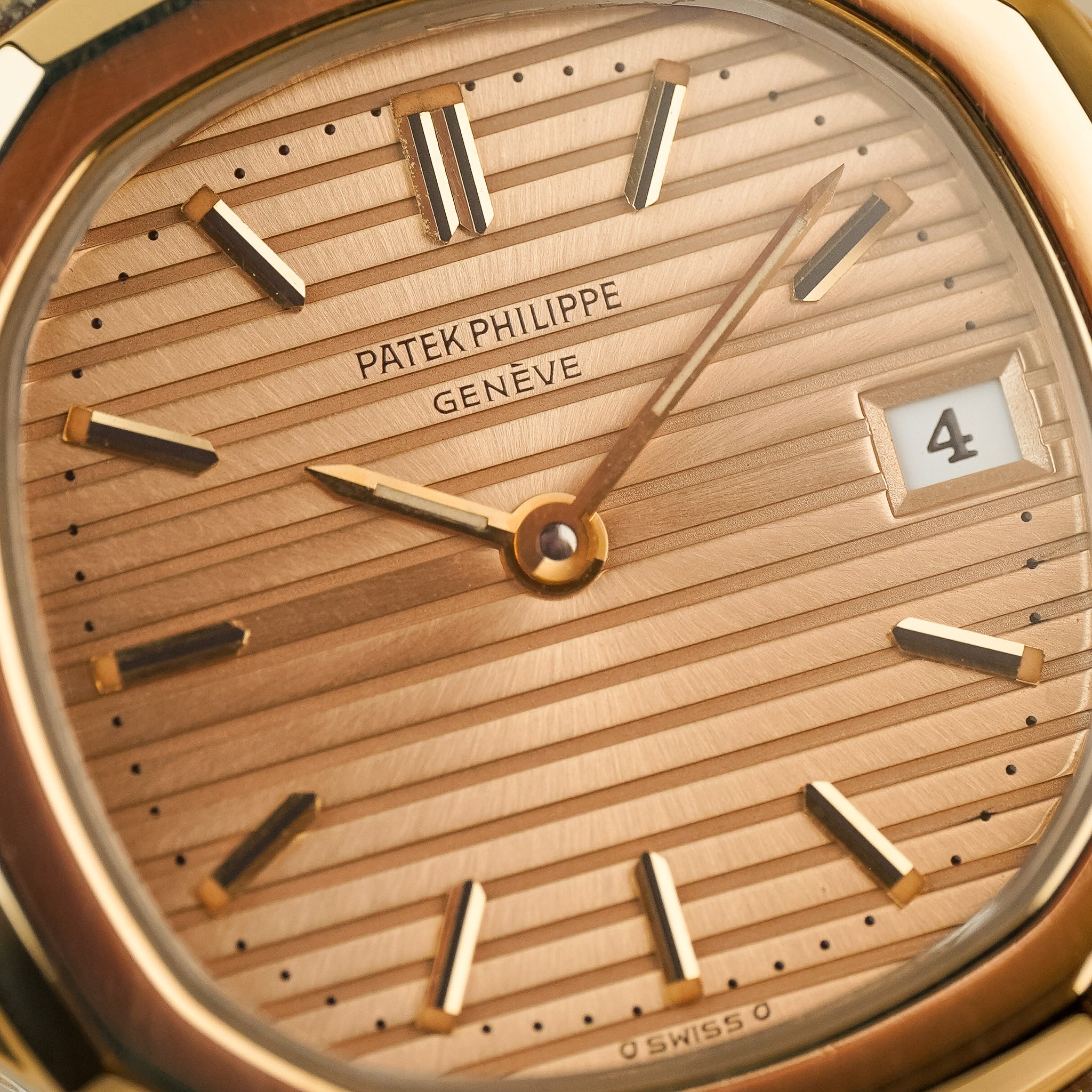 Close-up of a Patek Philippe Geneve wristwatch dial with a warm gold striped sunburst finish, gold-tone hands and hour markers, and a date window showing 4.