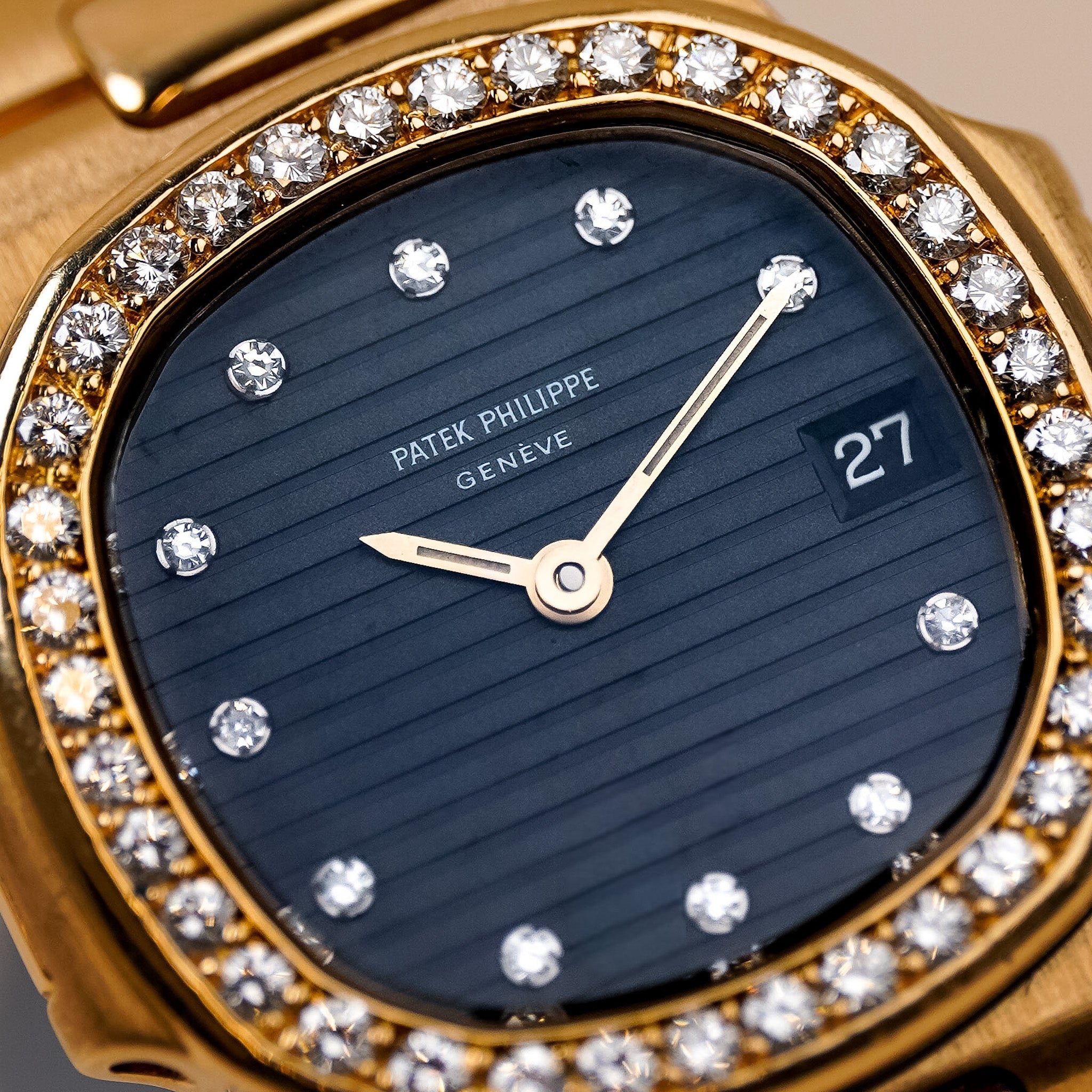 Closeup of a yellow gold Patek Philippe ladies watch with a blue dial, diamond bezel, diamond hour markers, and a date window showing "27"