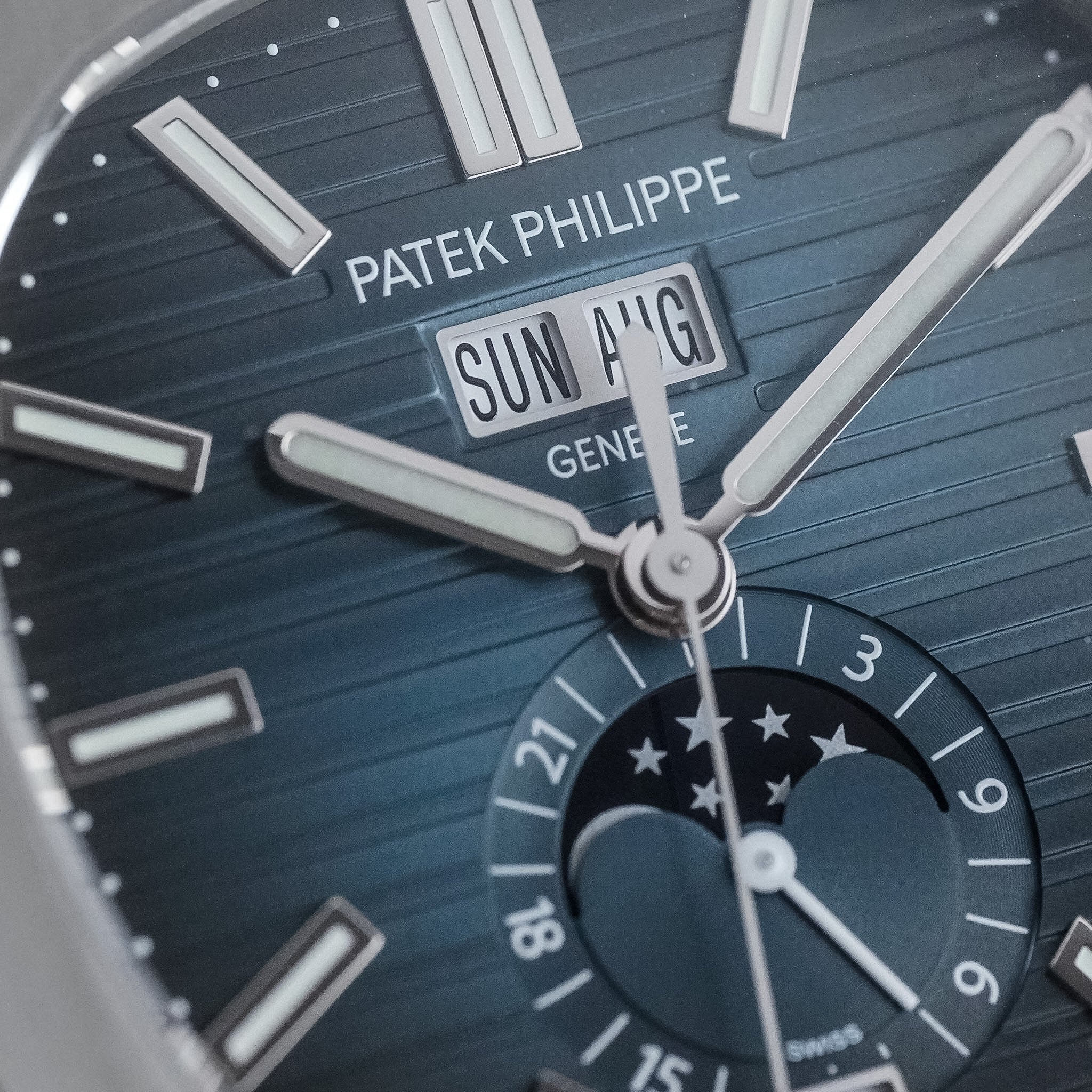 Close-up of a Patek Philippe wristwatch dial with a dark blue sunburst finish, silver-tone baton indices, a day-and-date window reading SUN ANG, and a moonphase subdial at the lower right.