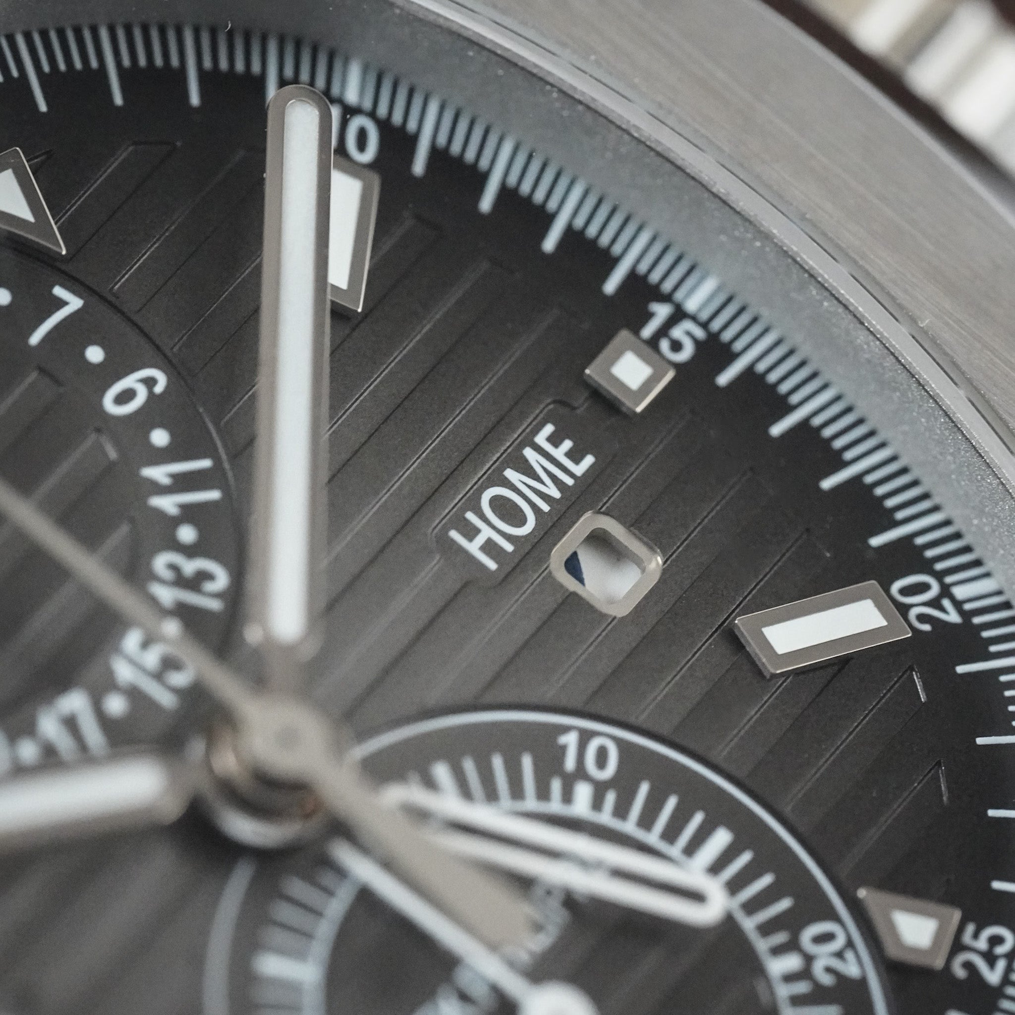 Close-up of a wristwatch dial with a dark gray or black textured bezel, silver-tone hands and markers, and the word HOME visible on the subdial.