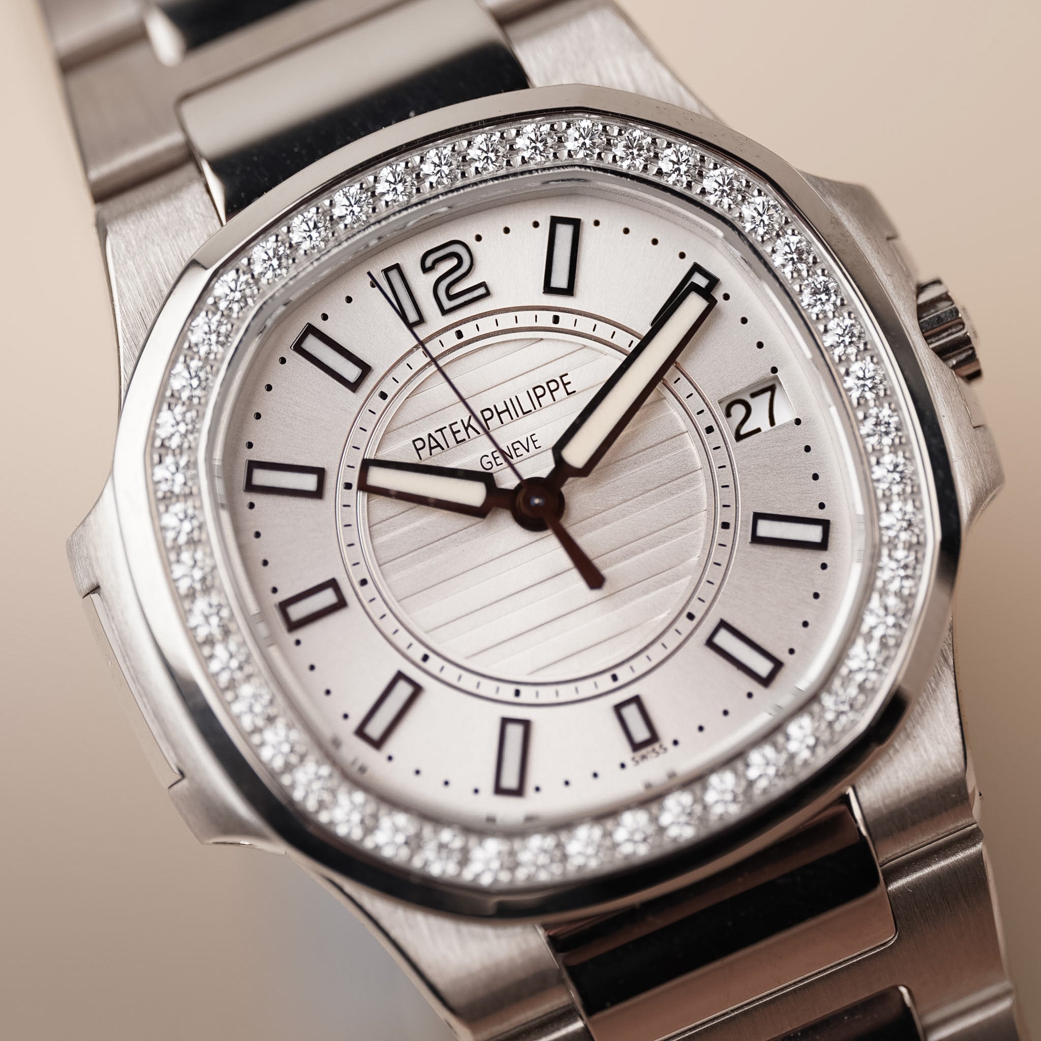 Closeup of a white gold ladies Patek Philippe Nautilus with a white dial, diamond bezel, and white gold bracelet. Shot laying flat against a solid beige surface.