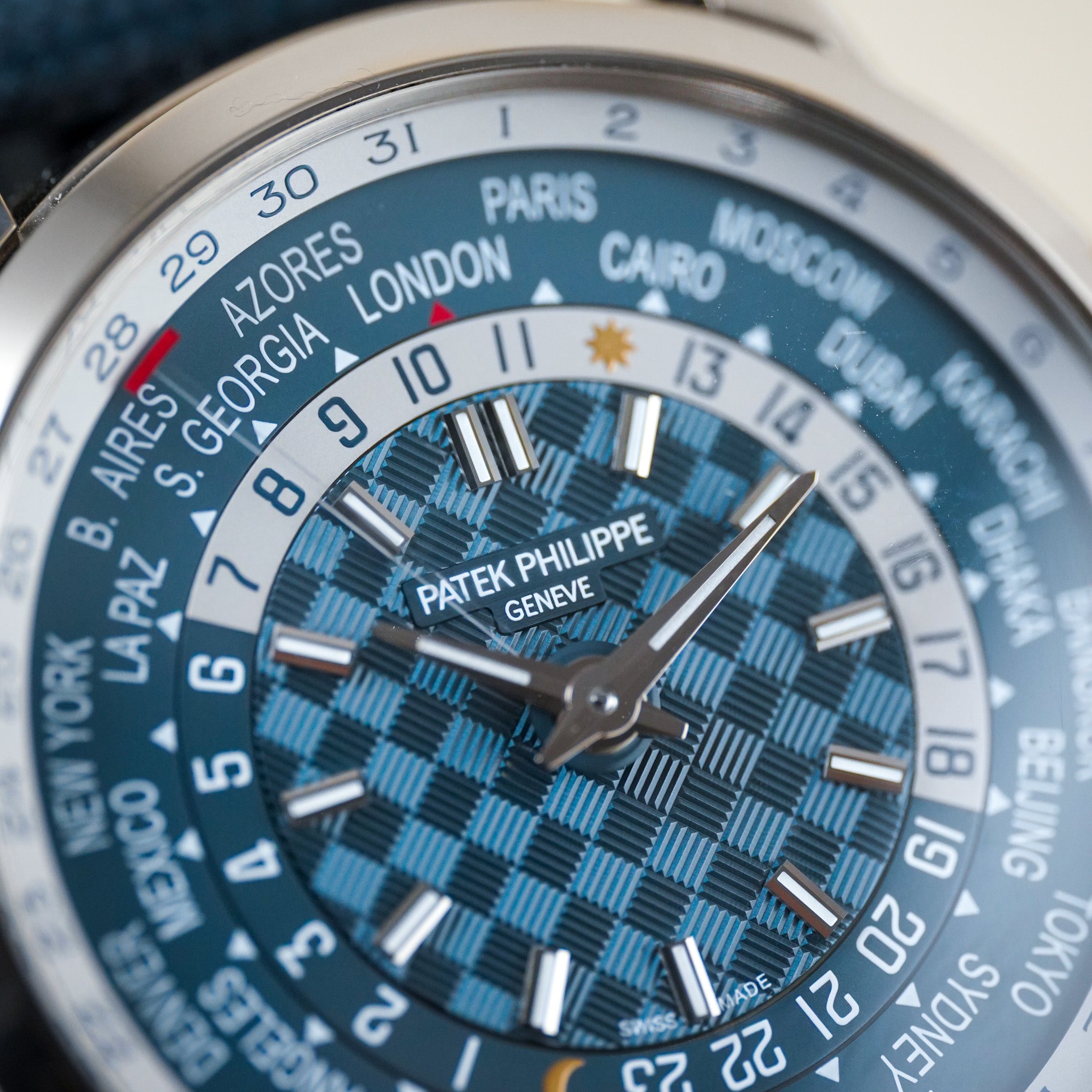 Close-up of a Patek Philippe wristwatch dial with a blue and black checkered pattern, world city time indicators around the bezel, and silver-tone hands and hour markers.