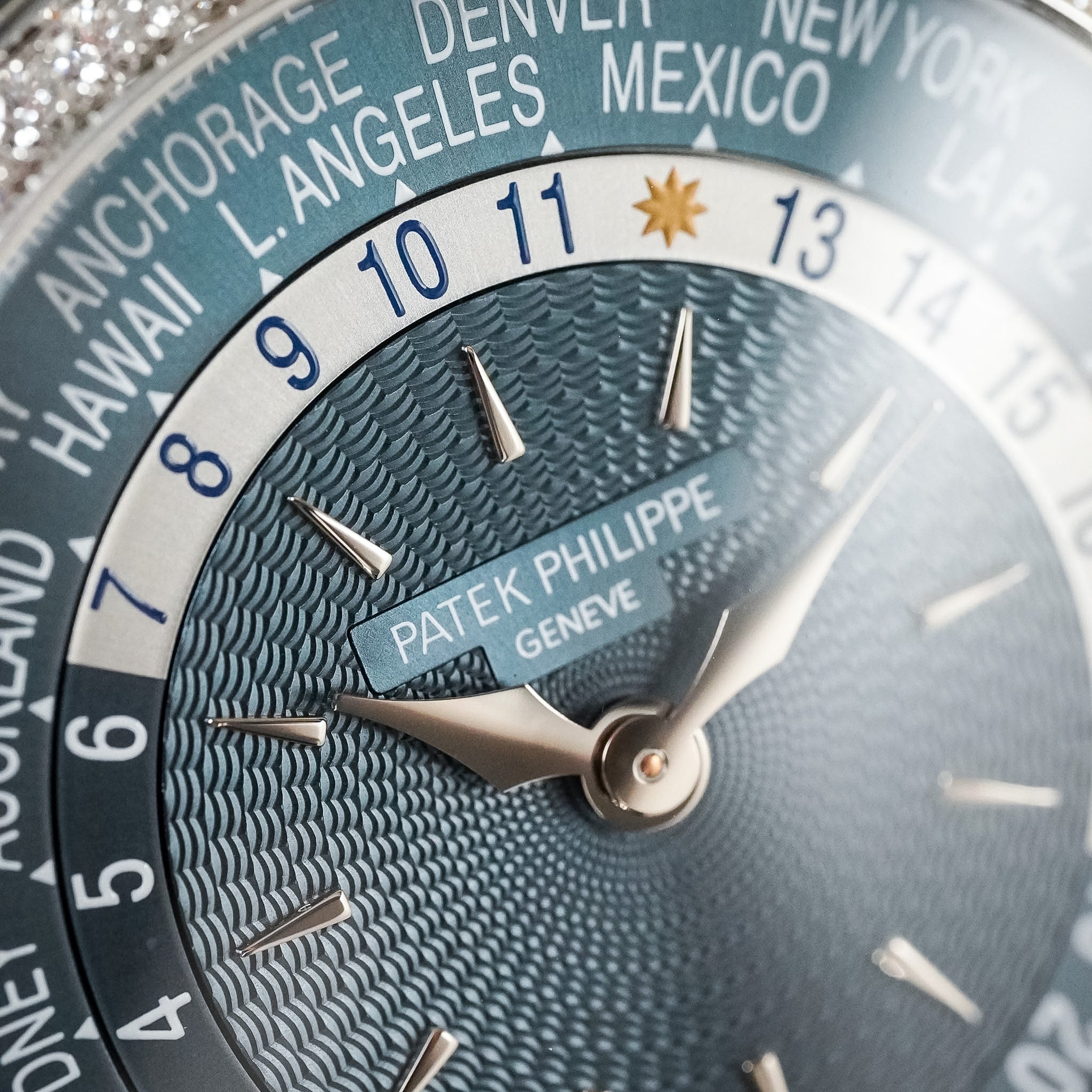 Close-up of a Patek Philippe Geneva world time watch dial, showing a textured blue wave pattern, silver hands, and a white ring with city labels including New York, Mexico, and Hawaii.