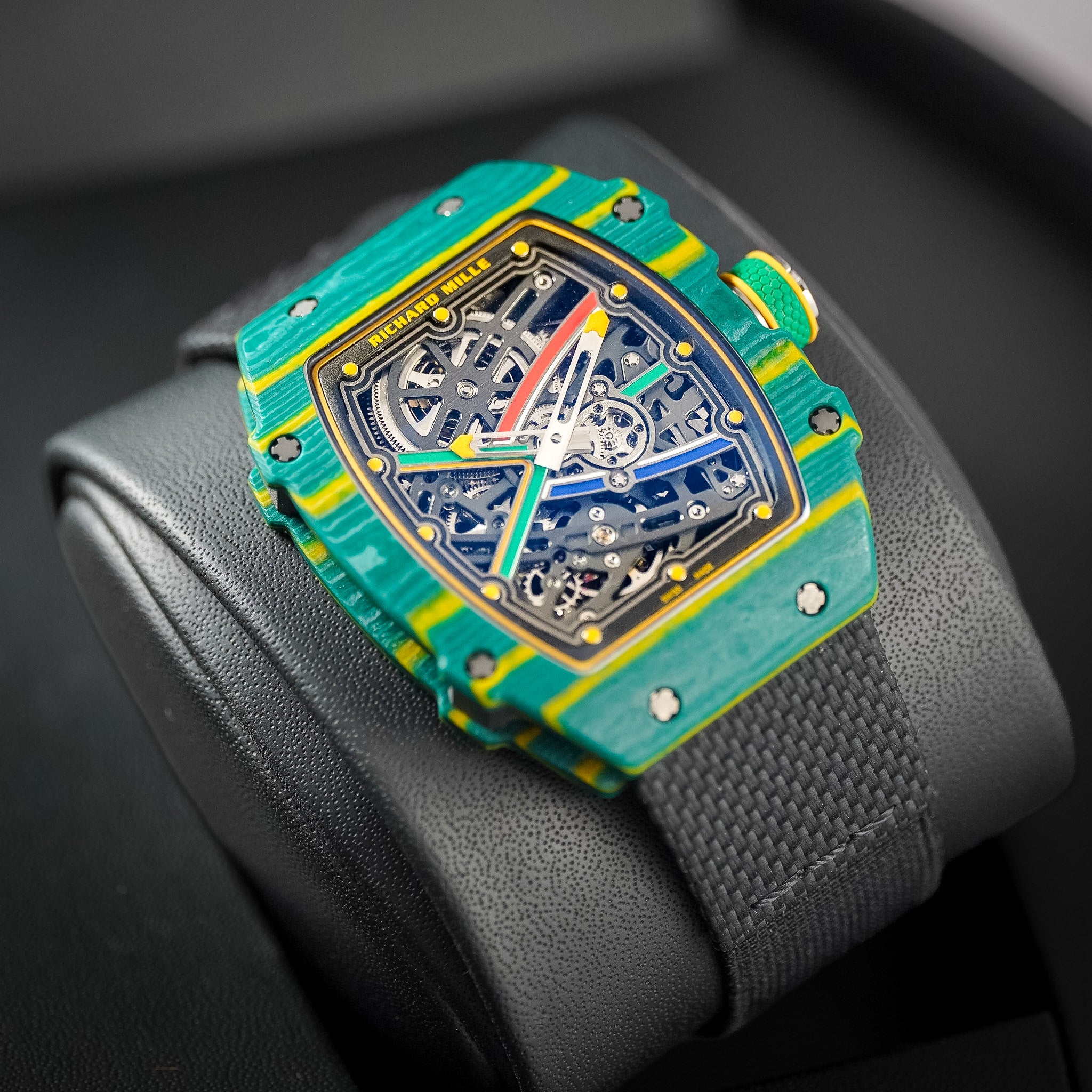 A Richard Mille wristwatch with a vibrant green and yellow textured bezel, a skeletonized dial showing intricate gears, and a black textured strap, resting on a black leather surface.