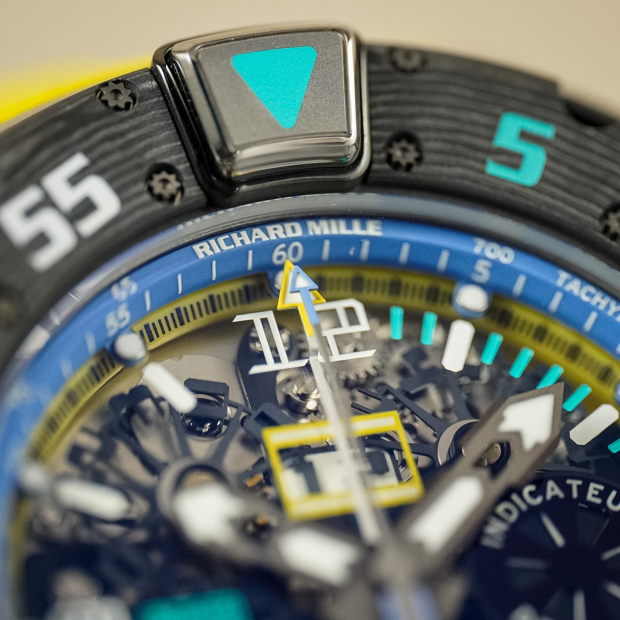 Close-up of a Richard Mille watch dial and bezel, showing a blue outer ring, black textured outer ring with white and turquoise markings, and a visible mechanical movement through a skeletonized case.