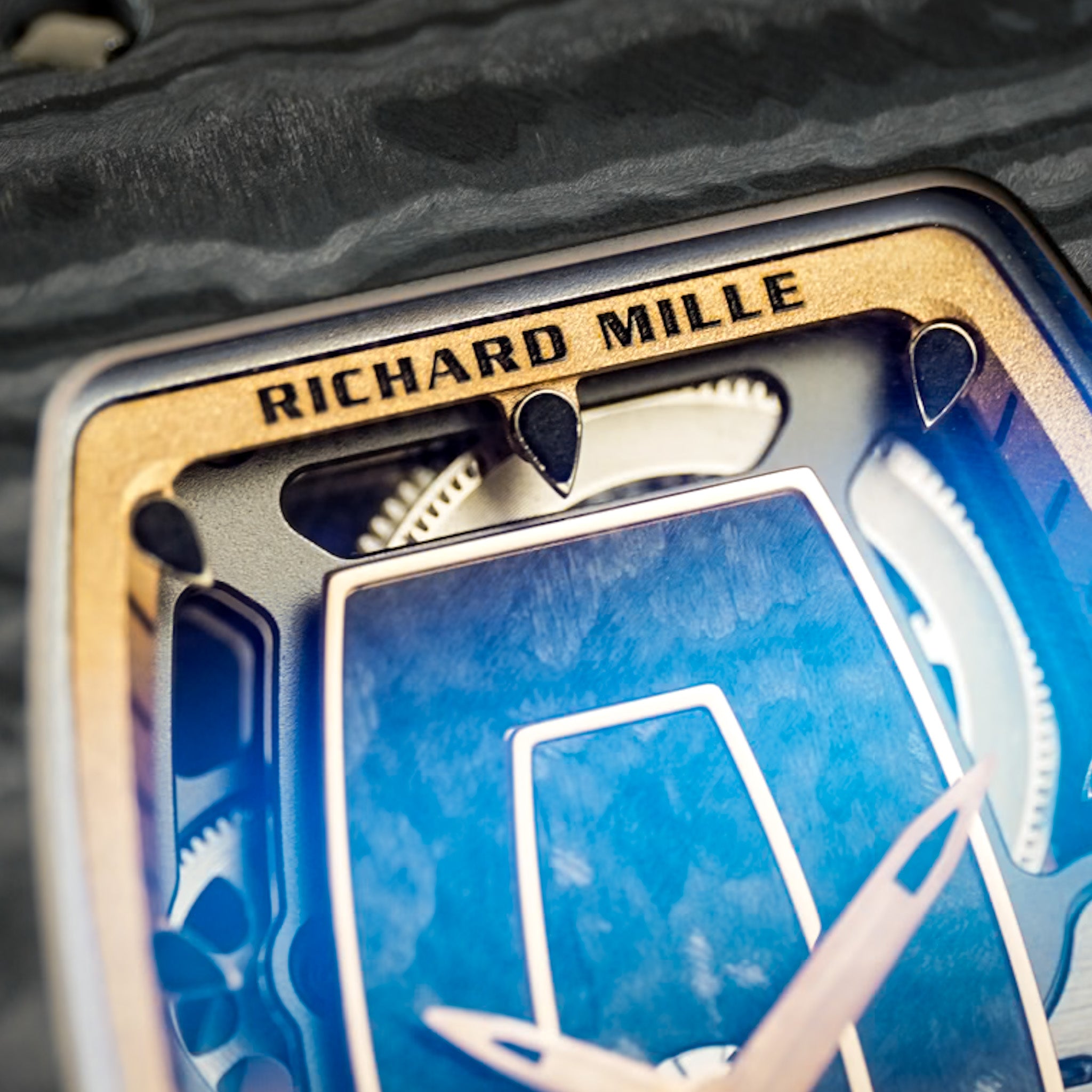 Close-up detail of a Richard Mille watch face with a blue dial, gold-toned bezel, and visible internal mechanical elements.