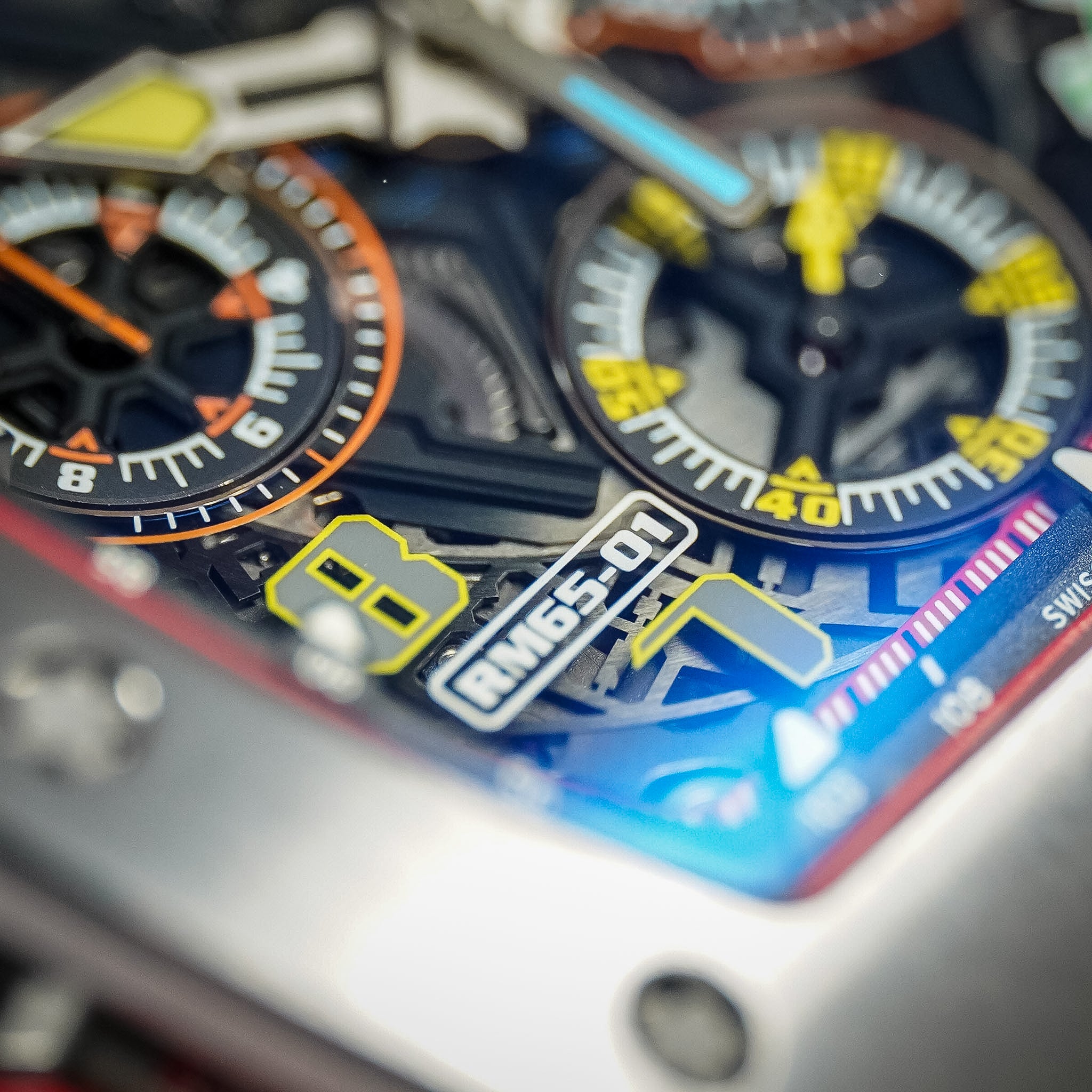 A closeup of a colorful skeletonized watch dial with a titanium case, RM65-01 logo, orange and yellow subdials, and a richly layered movement.