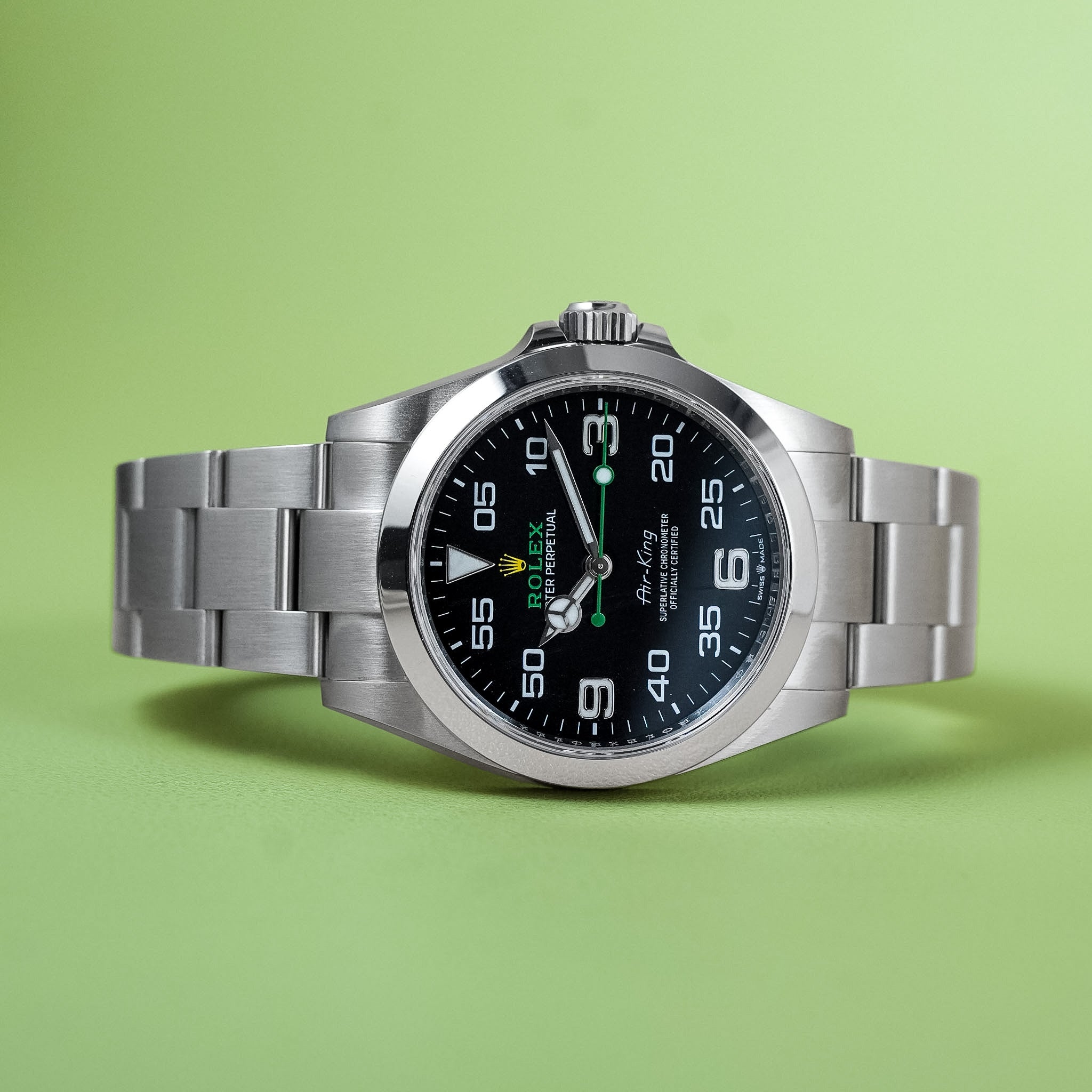 A Rolex Air-King watch with a stainless steel bracelet and black dial, photographed on a solid light green background