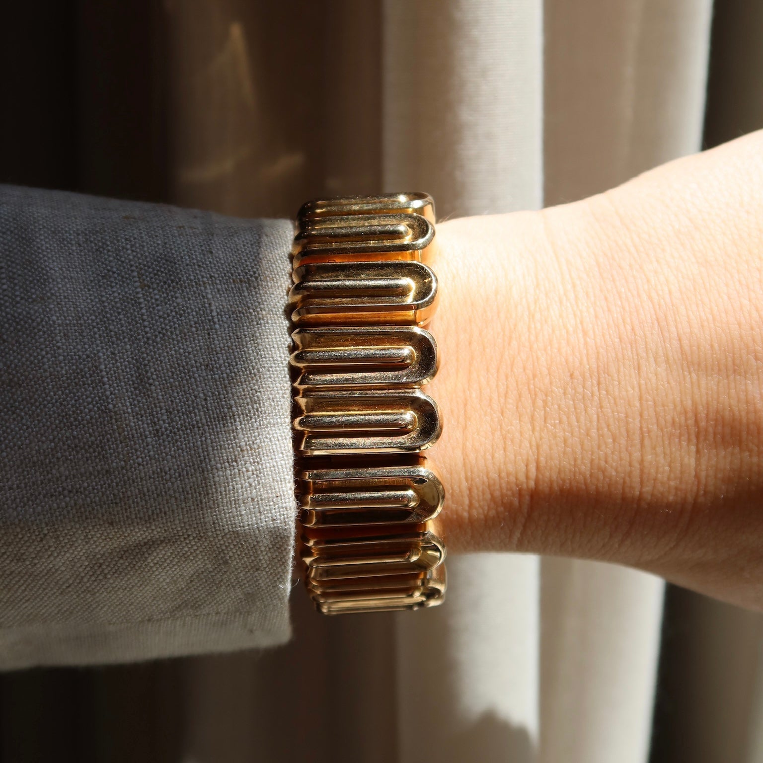 A woman's wrist wearing an art deco style rose gold bracelet in sunlight with a eige curtain background