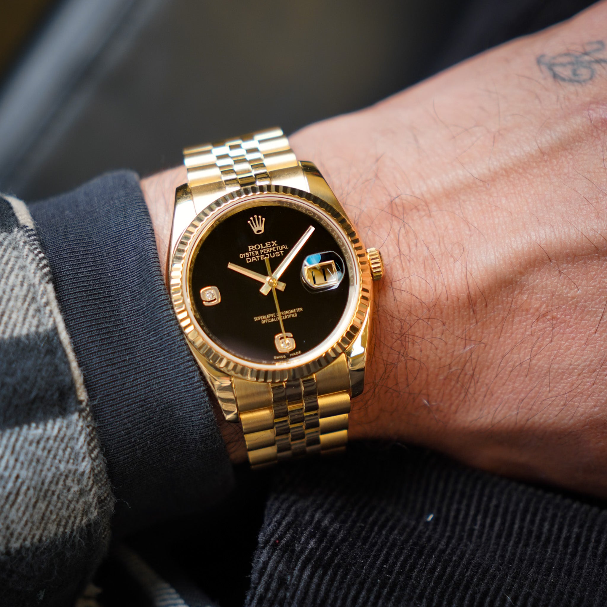 A man's wrist wearing a yellow gold Rolex Datejust with a fluted bezel, deep black onyx dial, gold Jubilee bracelet, and a date window showing "11". He is wearing a grey checkered outfit.