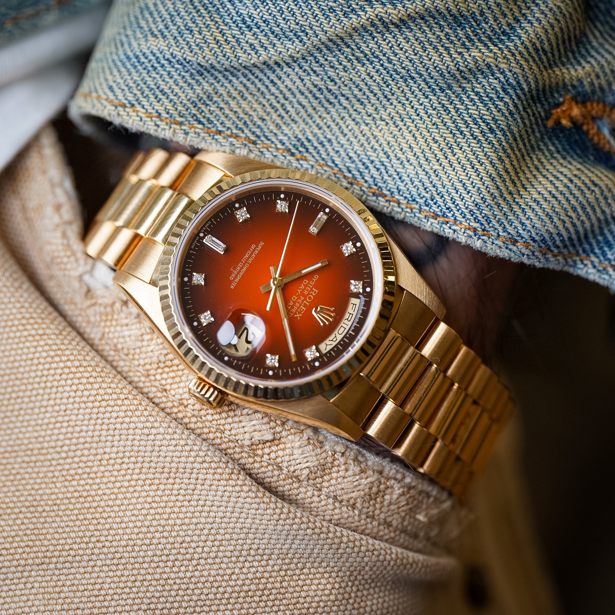 A yellow gold Rolex Day-Date watch with a burgundy ombré dial and diamond hour markers shot on a man's wrist wearing a denim jacket and tan pants